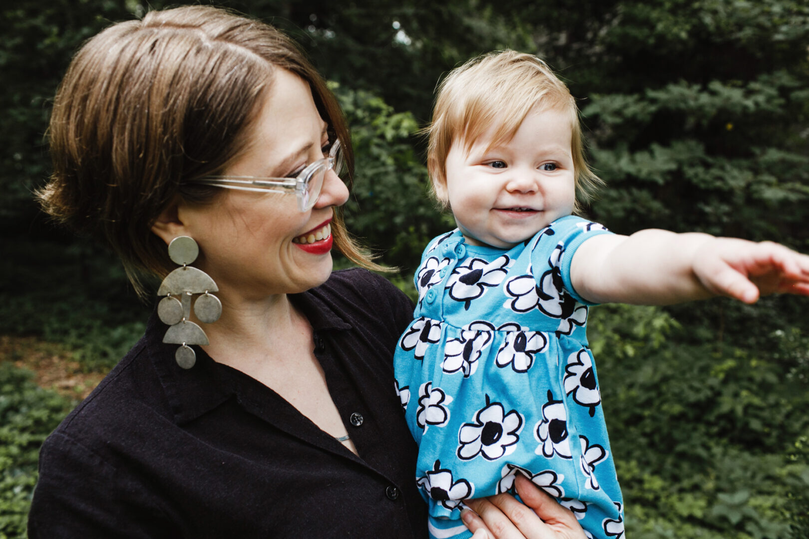 A woman holds a smiling baby in a blue floral dress, the little one pointing to the right. They are outdoors with trees in the background, capturing a precious moment perfect for their Philadelphia family photos.