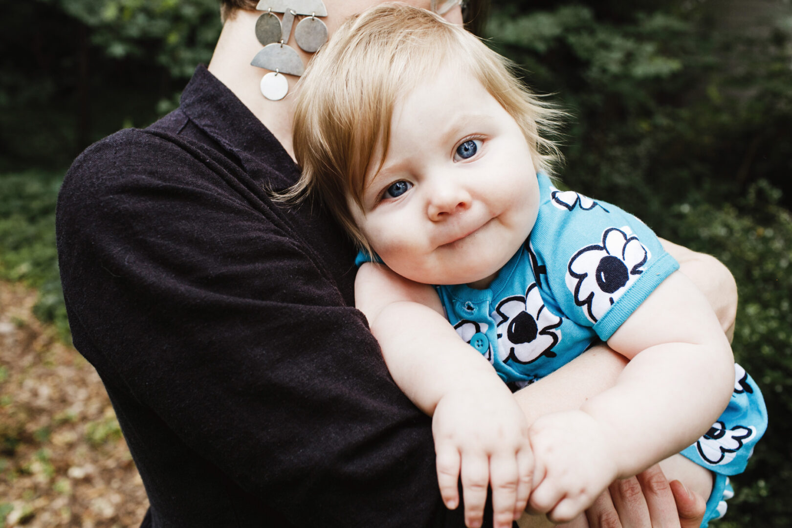 A baby with blue eyes and a floral blue outfit is cradled by a person with short hair in a dark shirt, capturing the essence of Philadelphia family photos.