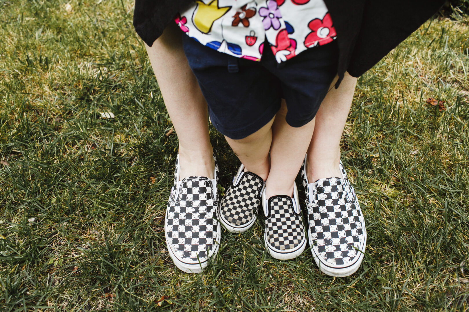 A child stands on an adult's feet, both wearing matching checkered slip-on shoes, creating a heartwarming moment captured perfectly for Philadelphia family photos on the grass.