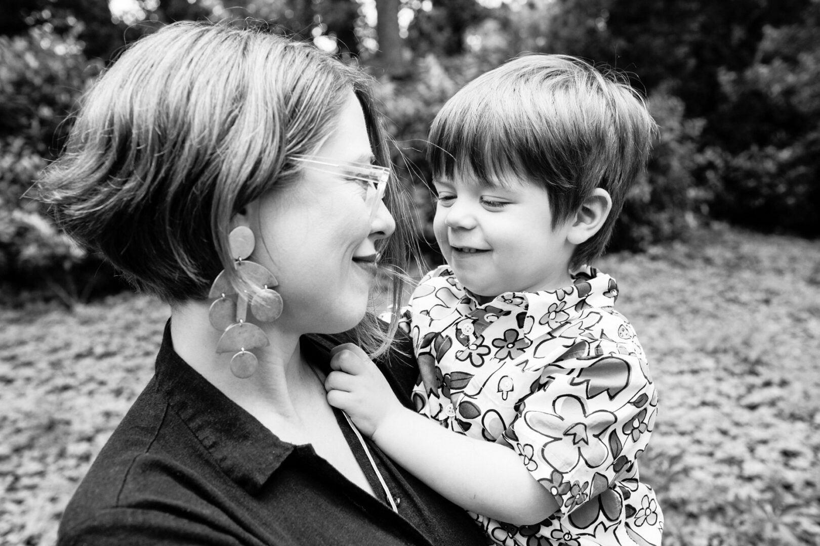 A black and white photo captures a joyful moment of a woman with short hair cradling a smiling child in a floral shirt, set against the lush backdrop of trees—an exquisite snapshot of cherished Philadelphia family photos.