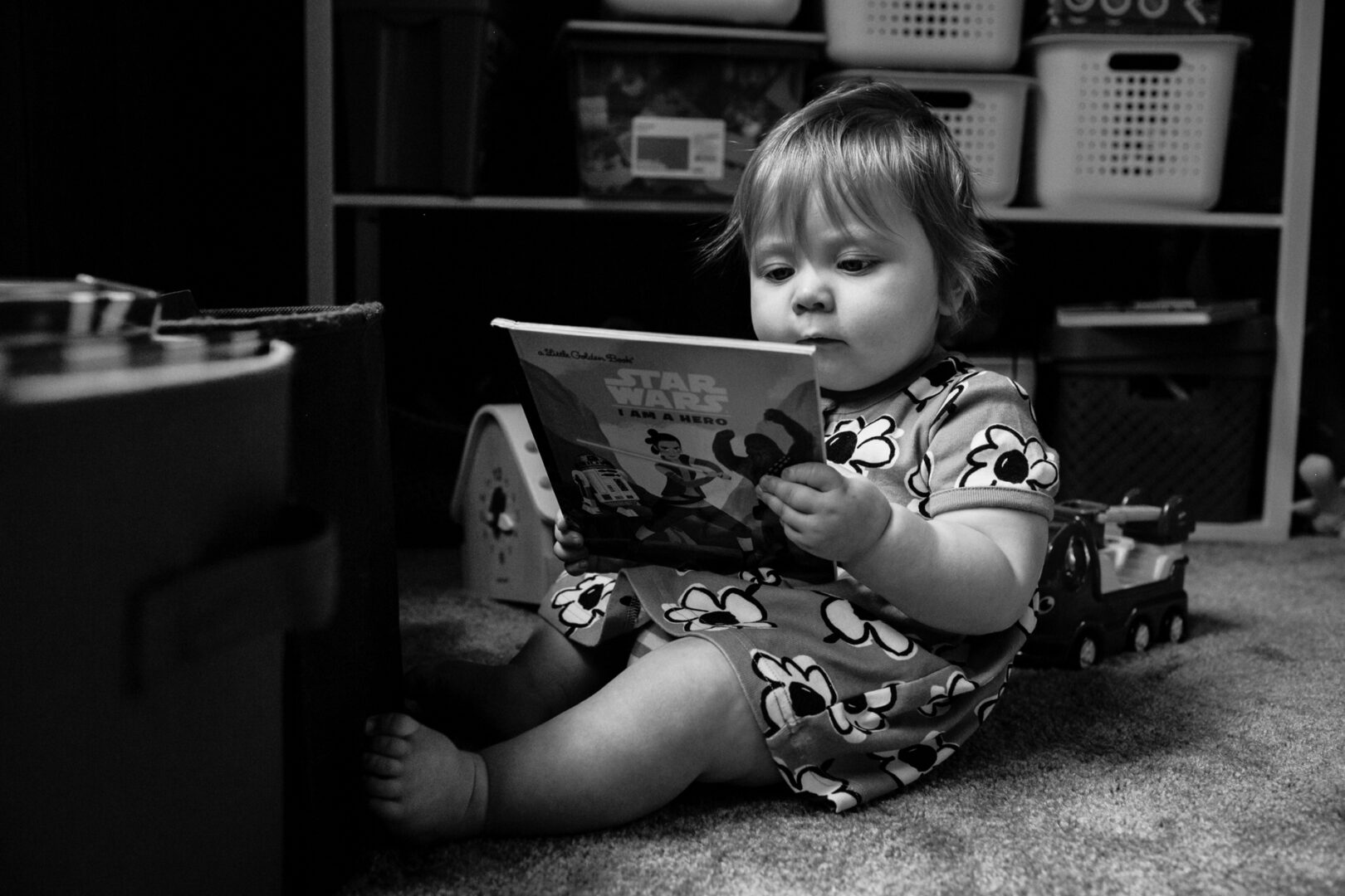 A black and white photo captures a toddler in Philadelphia, sitting on the floor engrossed in a "Star Wars" book, surrounded by toys and storage bins.