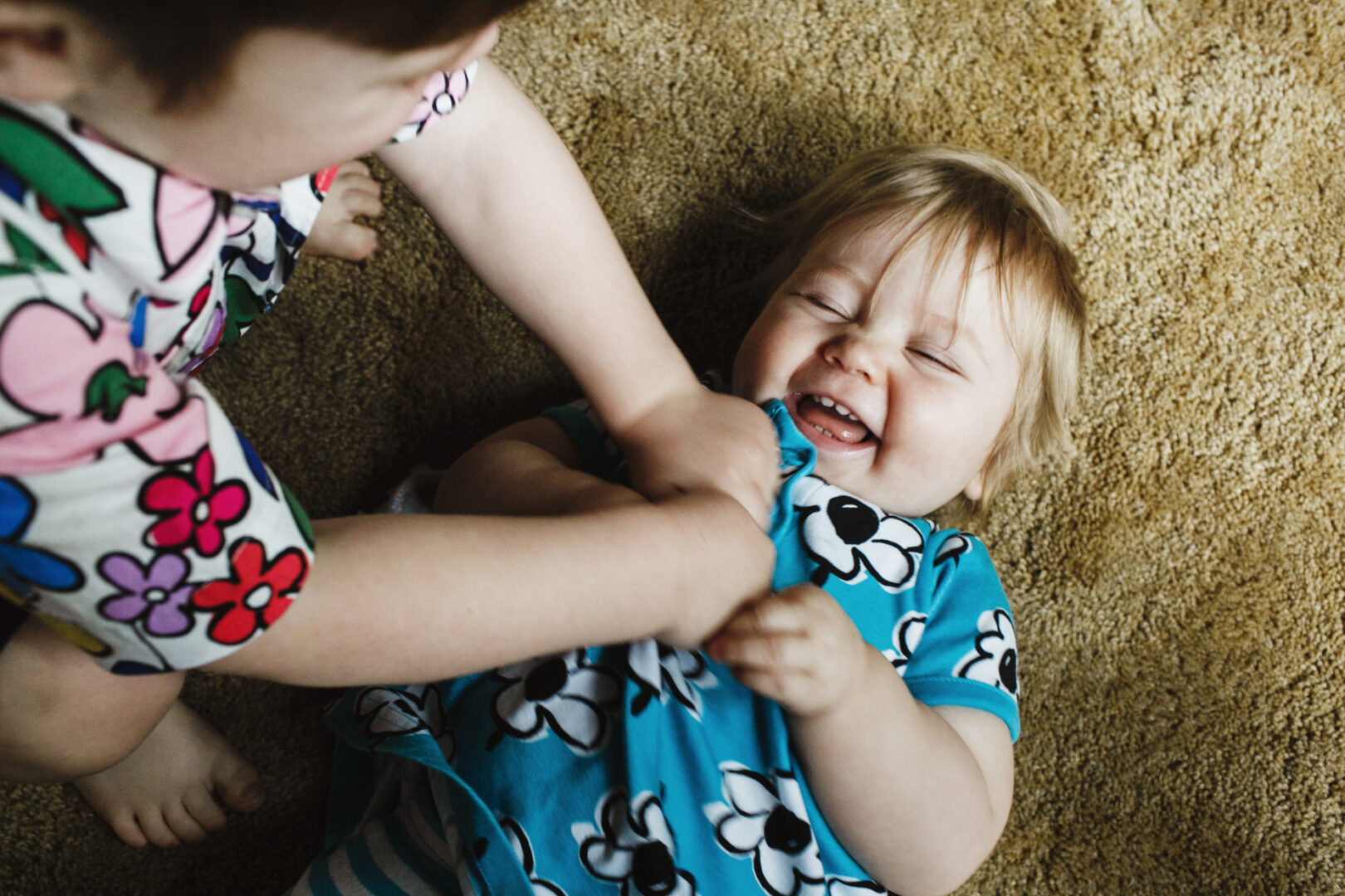 Two young children are on a carpeted floor in their cozy Philadelphia home; one is playfully tickling the other. The child being tickled, dressed in a blue floral outfit, laughs with eyes closed—a perfect snapshot for the family photo album.