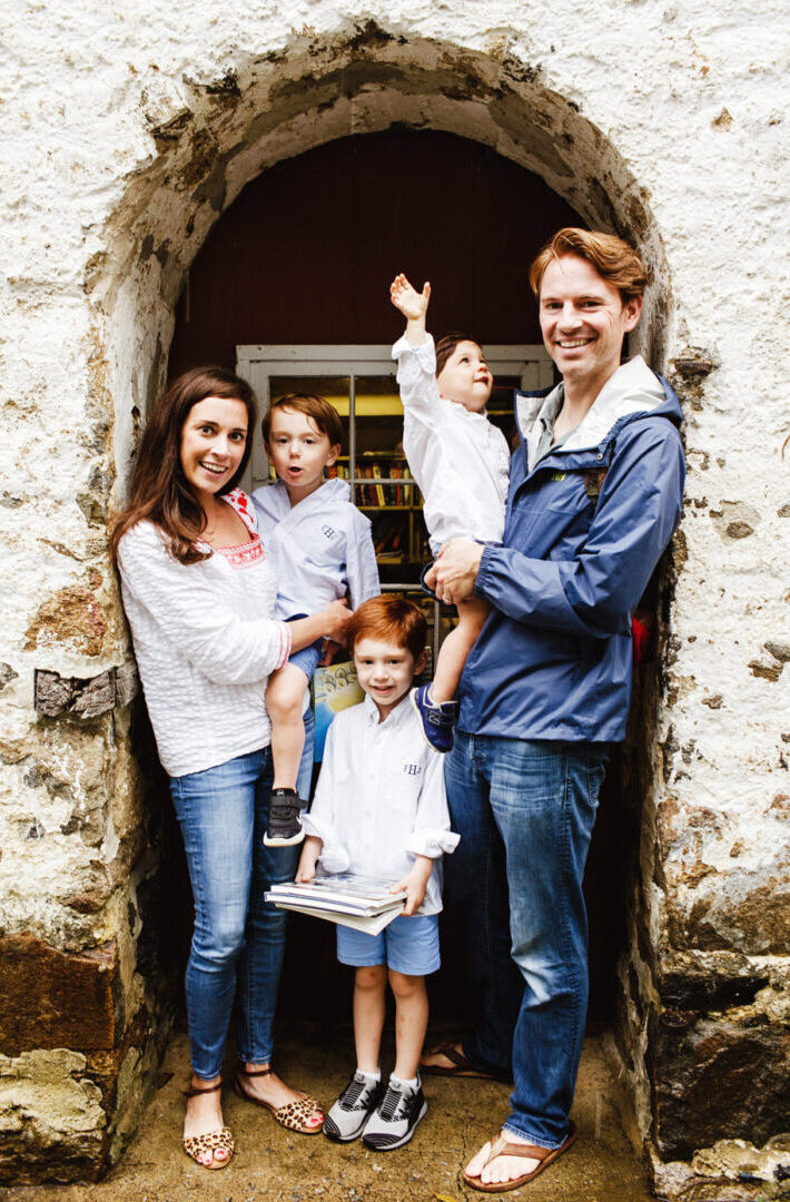 A West Chester family of five poses in front of a stone archway, exuding warmth. Parents hold two toddlers, while an older child stands proudly with books. Dressed in casual attire, their smiles capture the joy of cherished moments together.