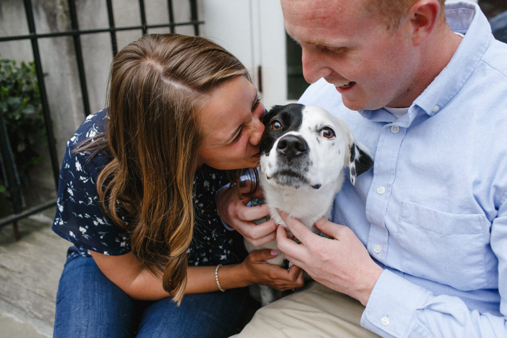 During their Chestnut Hill engagement session, a woman and a man sit on steps, smiling at a black and white dog. The woman kisses the dog, while the man gently holds it.