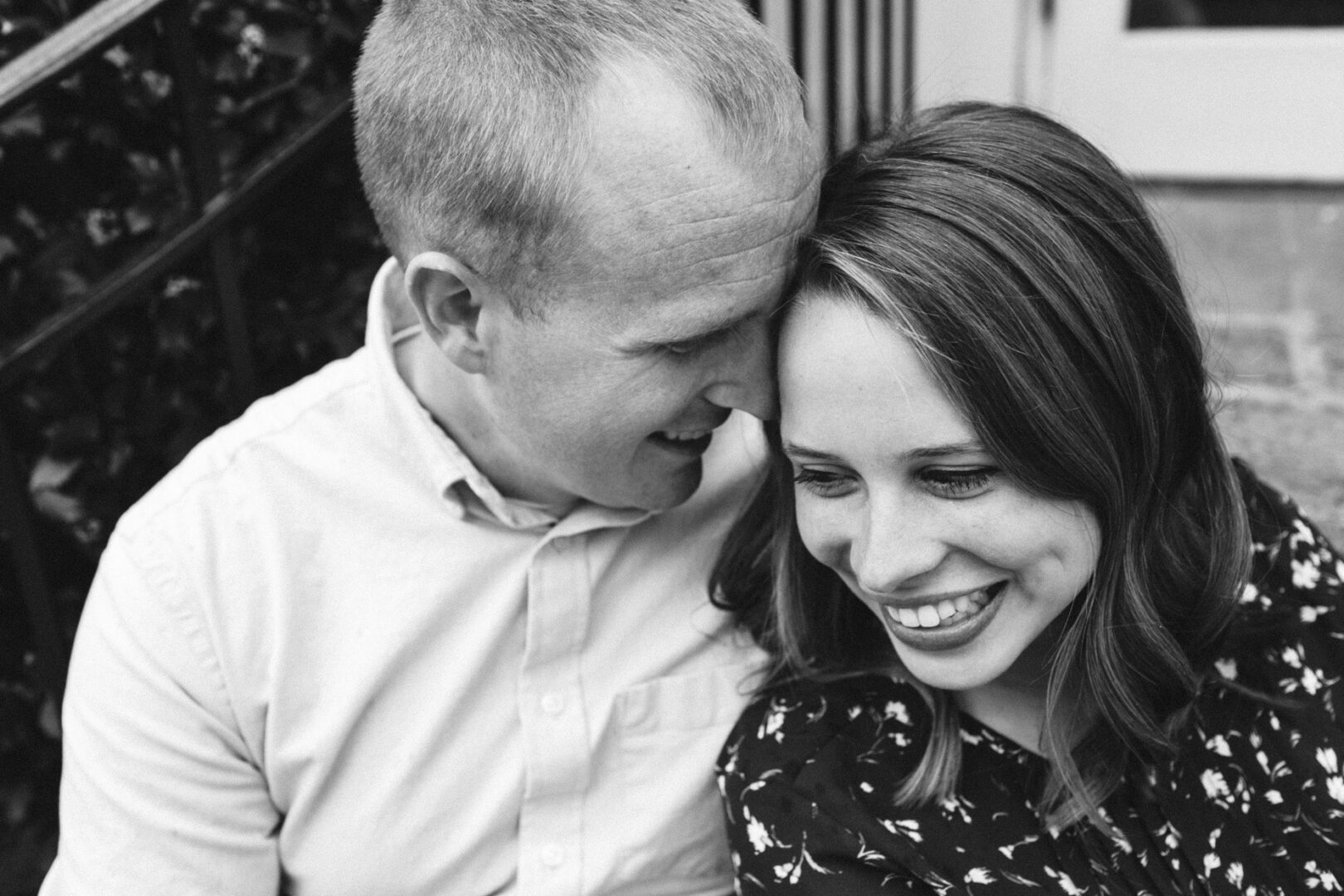 A couple sits closely on the stairs during their Chestnut Hill engagement session, the man whispering to the smiling woman. The black and white image captures a timeless moment of intimacy.