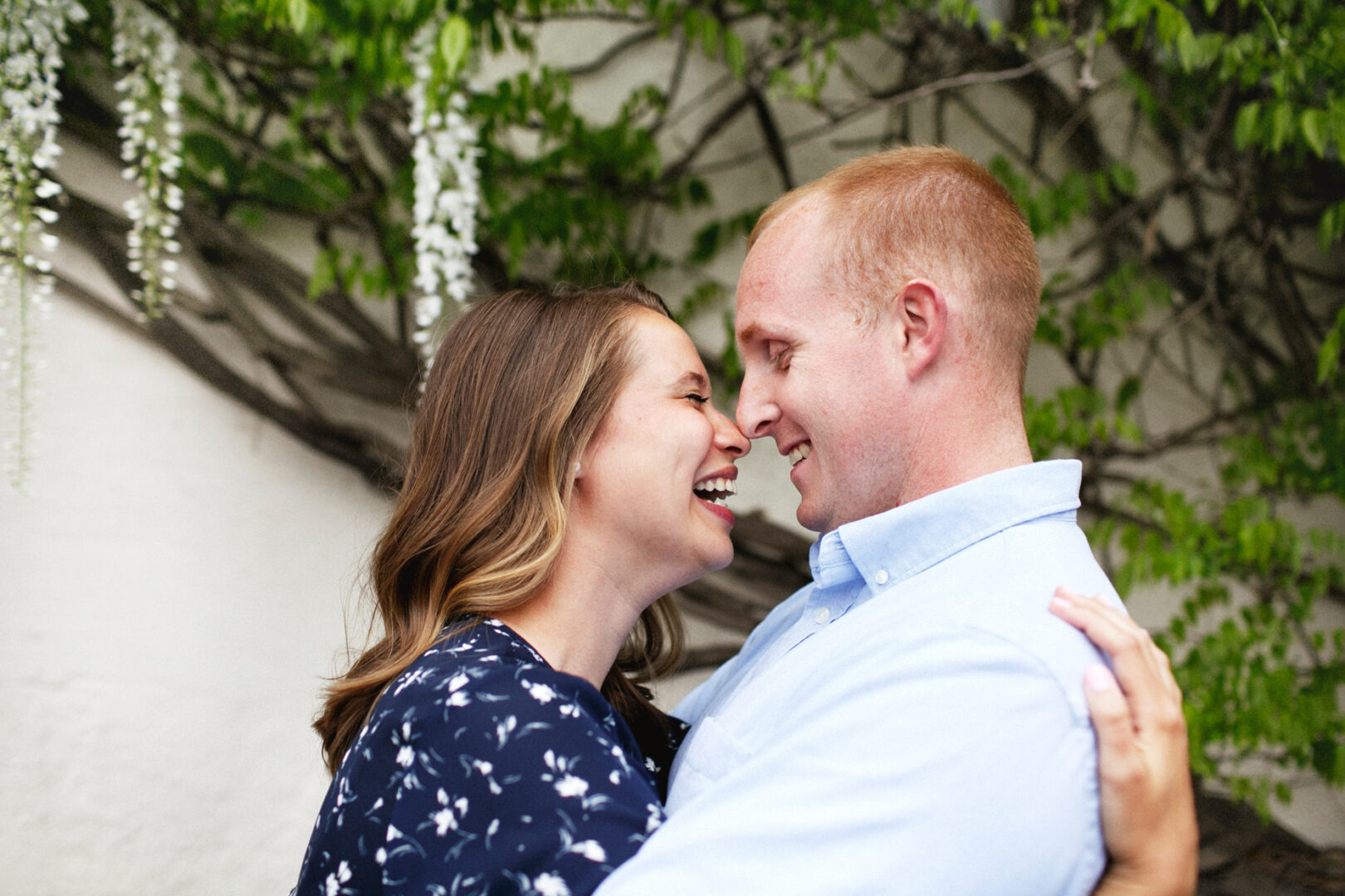 A couple smiling at each other, standing close together amidst a backdrop of leafy branches during their Chestnut Hill engagement session, with the woman in a patterned dress and the man clad in a light blue shirt.