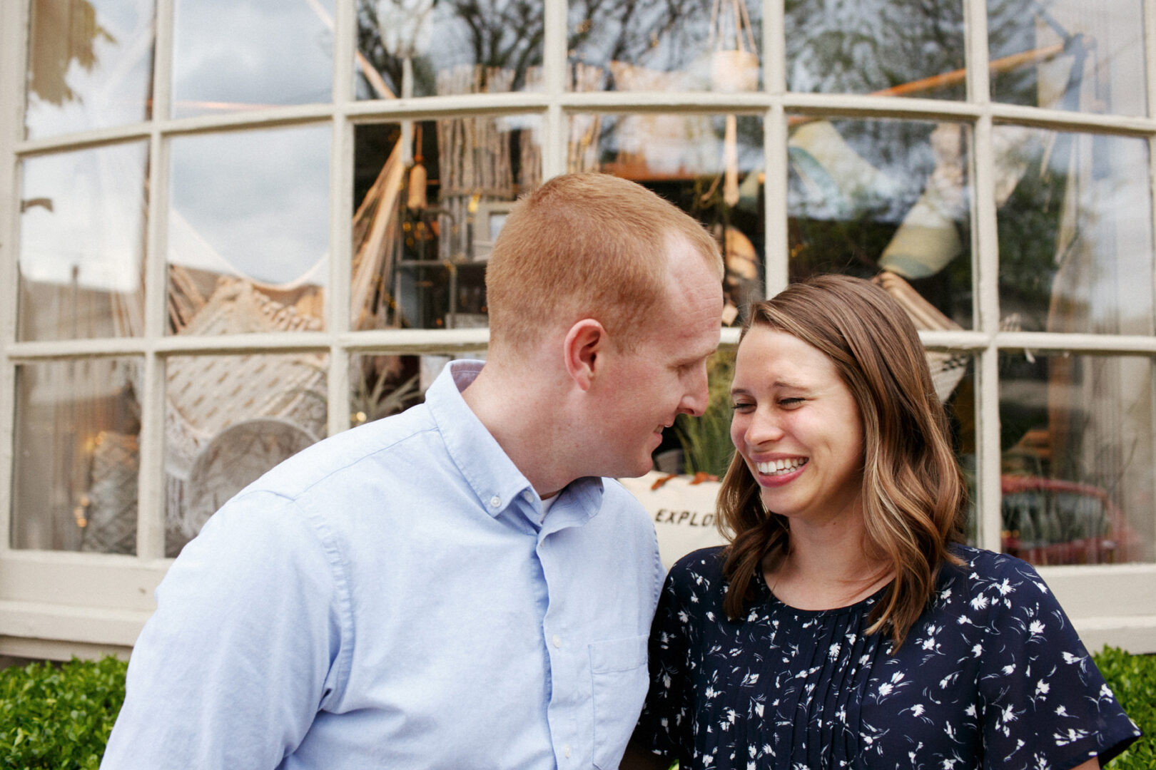 A couple stands in front of a large window, exchanging smiles during their Chestnut Hill engagement session. The man wears a light blue shirt, while the woman dazzles in a dark floral dress, their reflections gently mirrored in the glass.