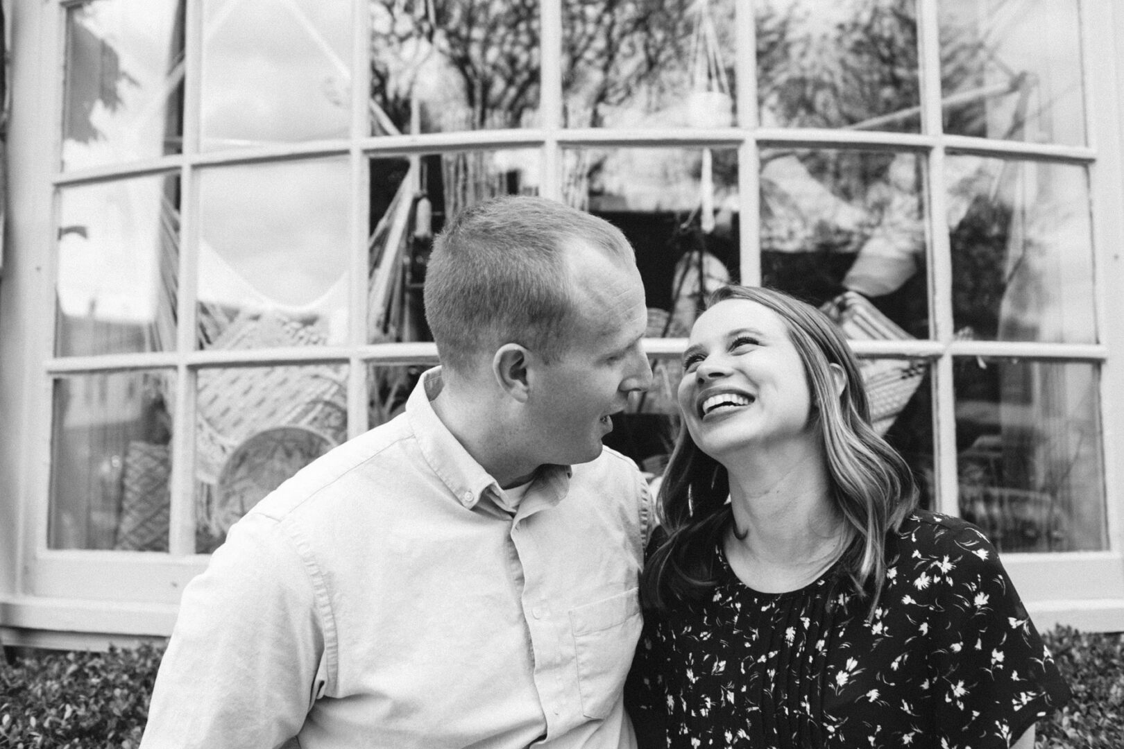 A man and a woman share a laugh in front of a large window with reflections of trees, capturing the joy of their Chestnut Hill engagement session. Black and white image.