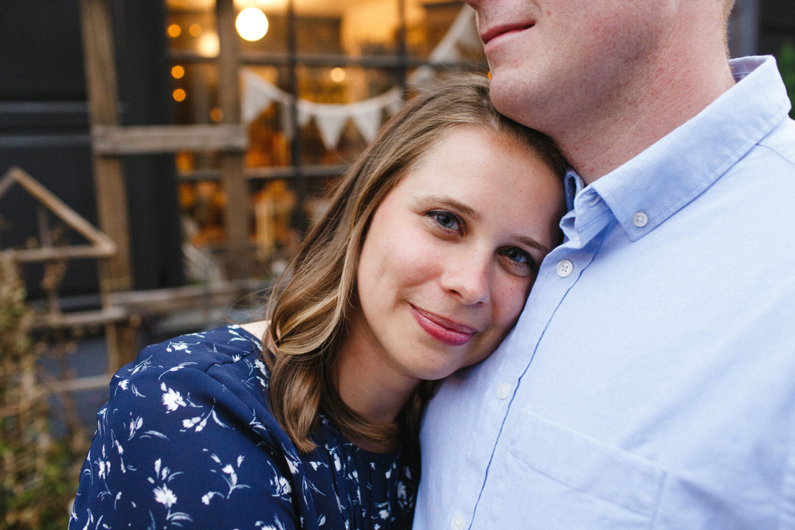 A woman smiles, resting her head on a man's chest, who wears a light blue shirt. They stand outdoors in front of a window display with decorative items, capturing the warmth of their Chestnut Hill engagement session.