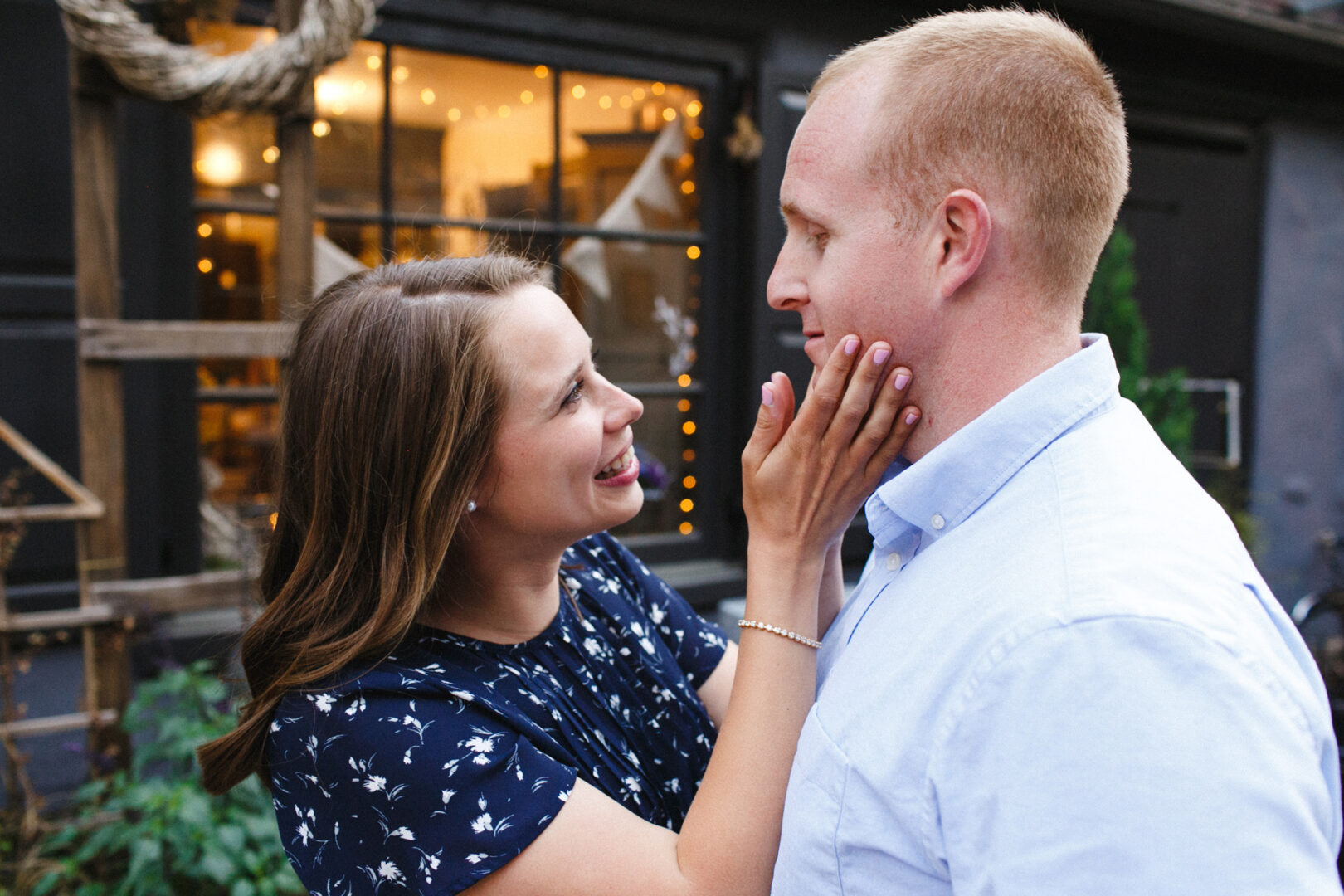 A woman in a blue floral dress smiles, gently touching a man's face. He, in a light blue shirt, gazes back at her lovingly. They stand outside near a window adorned with string lights, capturing the warmth of their Chestnut Hill Engagement Session.