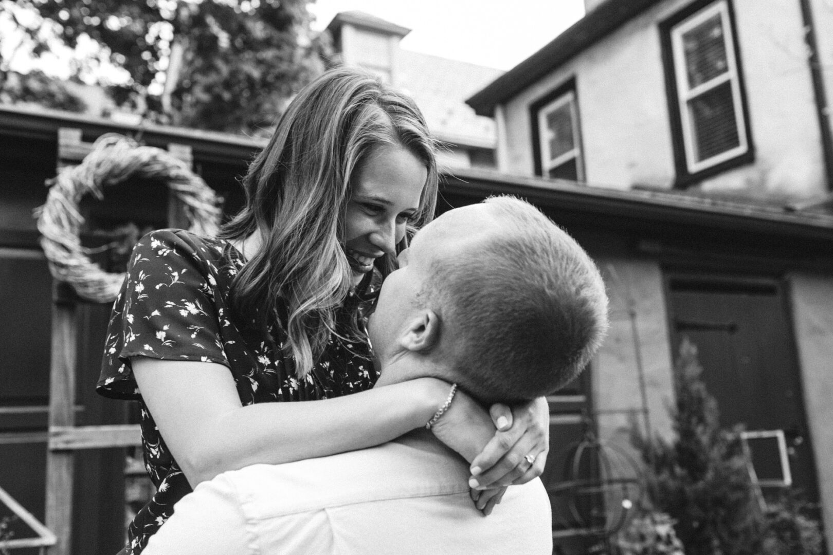 During a Chestnut Hill engagement session, a man lifts a smiling woman outdoors as they gaze at each other affectionately. In the background, a house adorned with a wreath adds charm to this timeless black and white image.