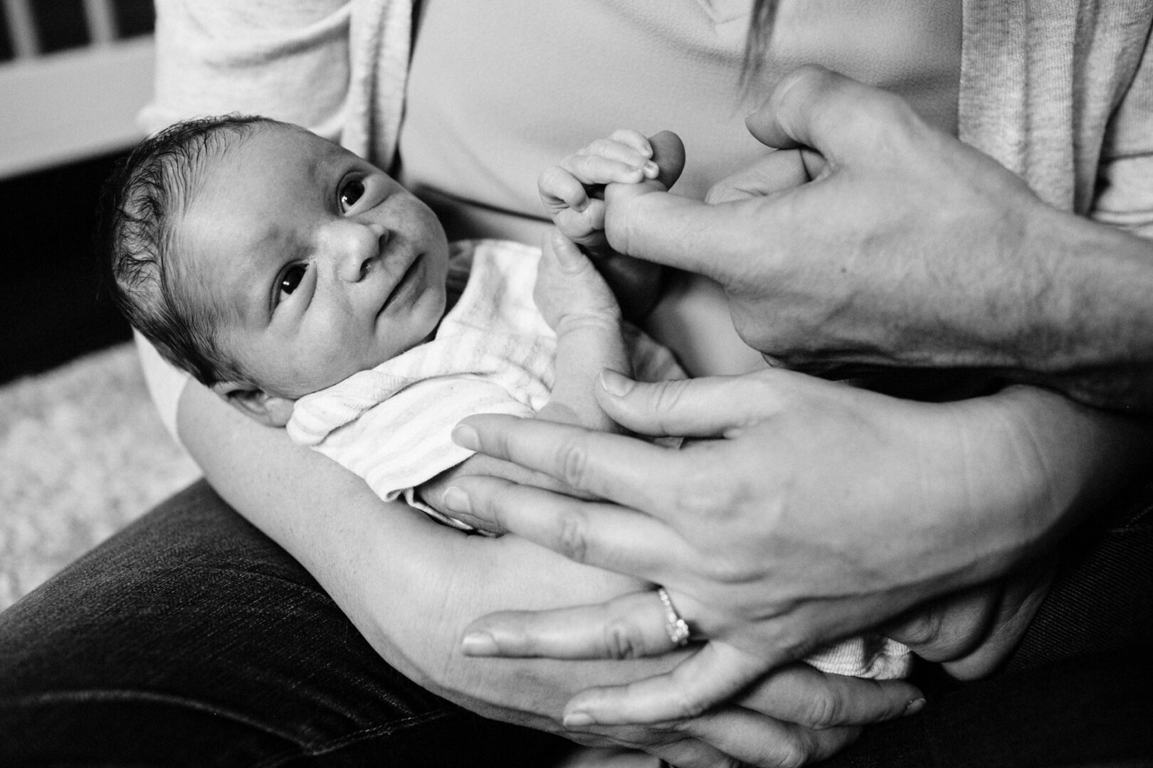 A newborn baby holds an adult's finger while being cradled in another person's arms, capturing the tender moment of newborn photos at home. The image is black and white.