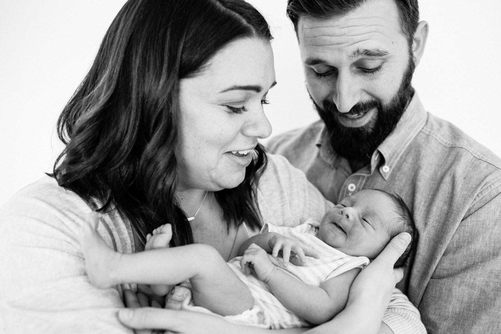 A couple in casual clothes holds their newborn baby at home, gazing at the infant with joy and affection. The image, captured in black and white, beautifully encapsulates these precious first moments.