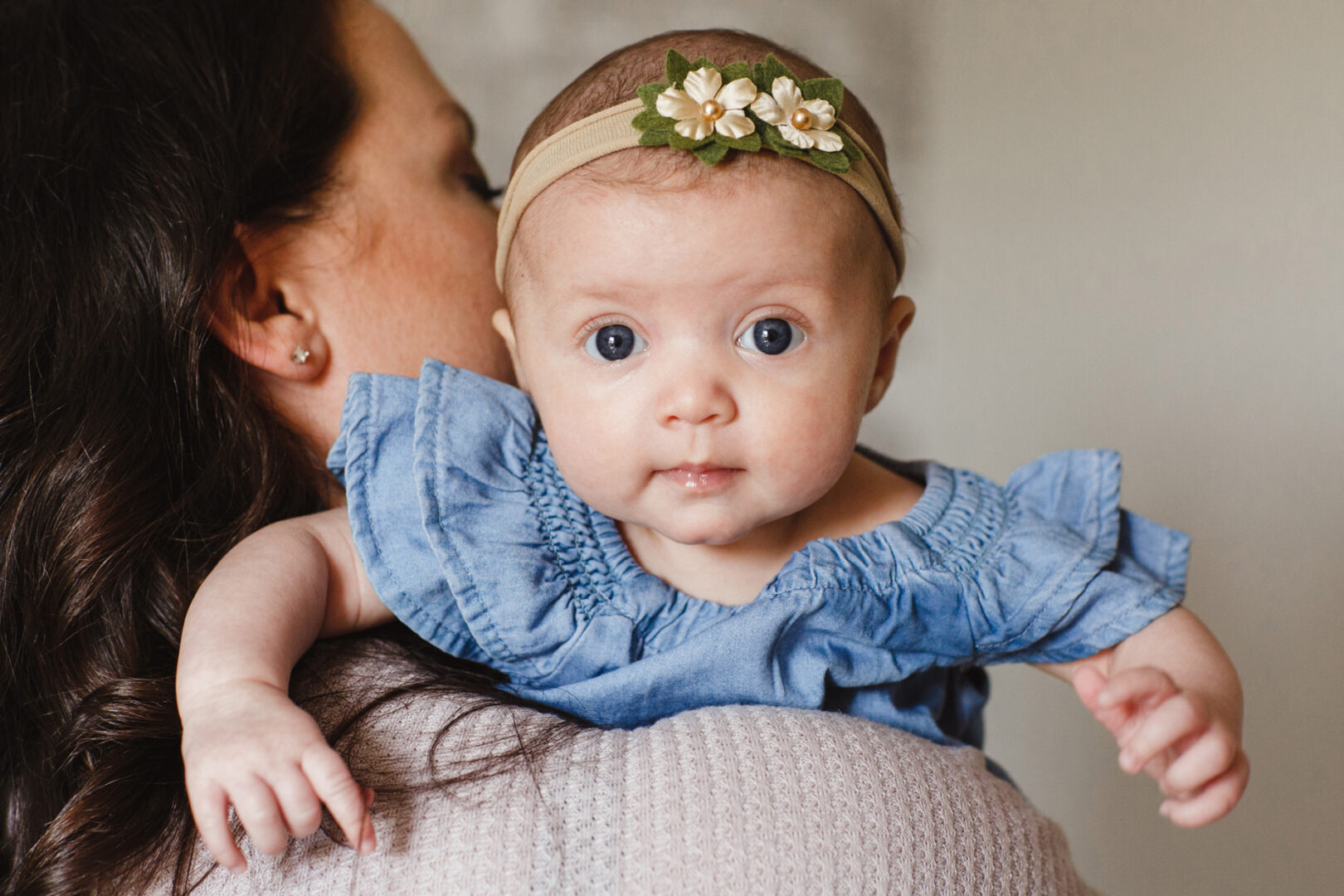 A baby in a blue dress and floral headband is gently held by an adult, gazing forward, capturing the essence of serene newborn photos in Philadelphia.
