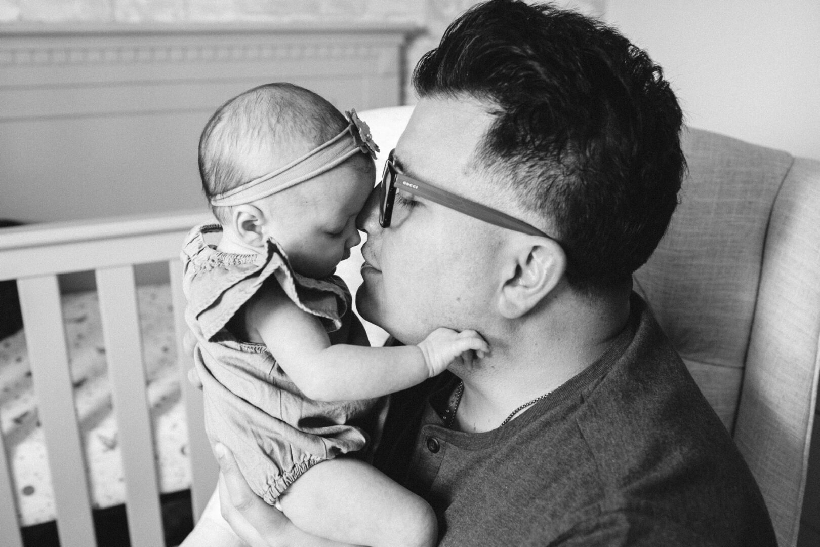 A man tenderly holds a baby close to his face in an affectionate gesture, seated near a crib. The newborn, donned in a headband and dress, is the focus of this timeless black and white image. Captured with love, it's the essence of newborn photos in Philadelphia.