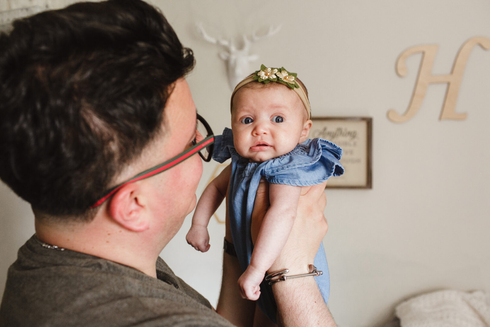 An adult cradles a baby in a blue outfit adorned with a floral headband, capturing a tender moment. This beautiful scene, perfect for newborn photos in Philadelphia, unfolds indoors, with decorative letters adding charm to the backdrop.