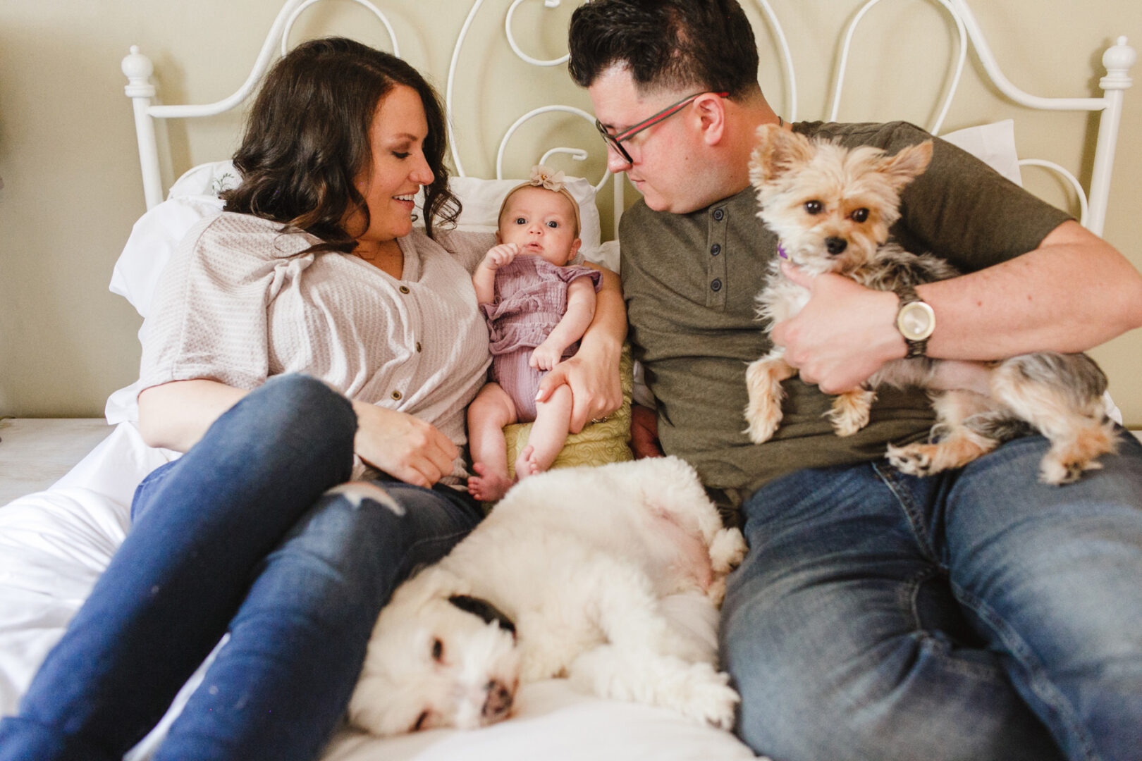 A couple sits on a bed with their newborn and two dogs. The woman smiles at the baby, while the man holds a small dog. A larger dog rests beside them, capturing a perfect moment of family bliss for these Philadelphia residents.