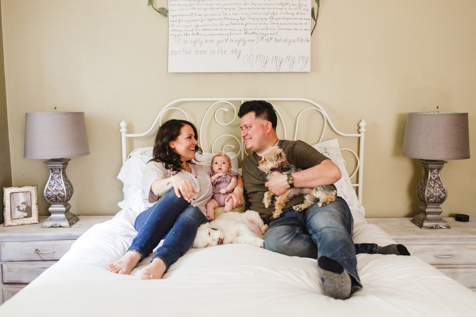 A couple sits on a bed with a newborn nestled between them, capturing a tender moment in their Philadelphia home. A small dog rests on the man's lap as a decorative sign hangs above the headboard, while two bedside tables with lamps flank this precious tableau.