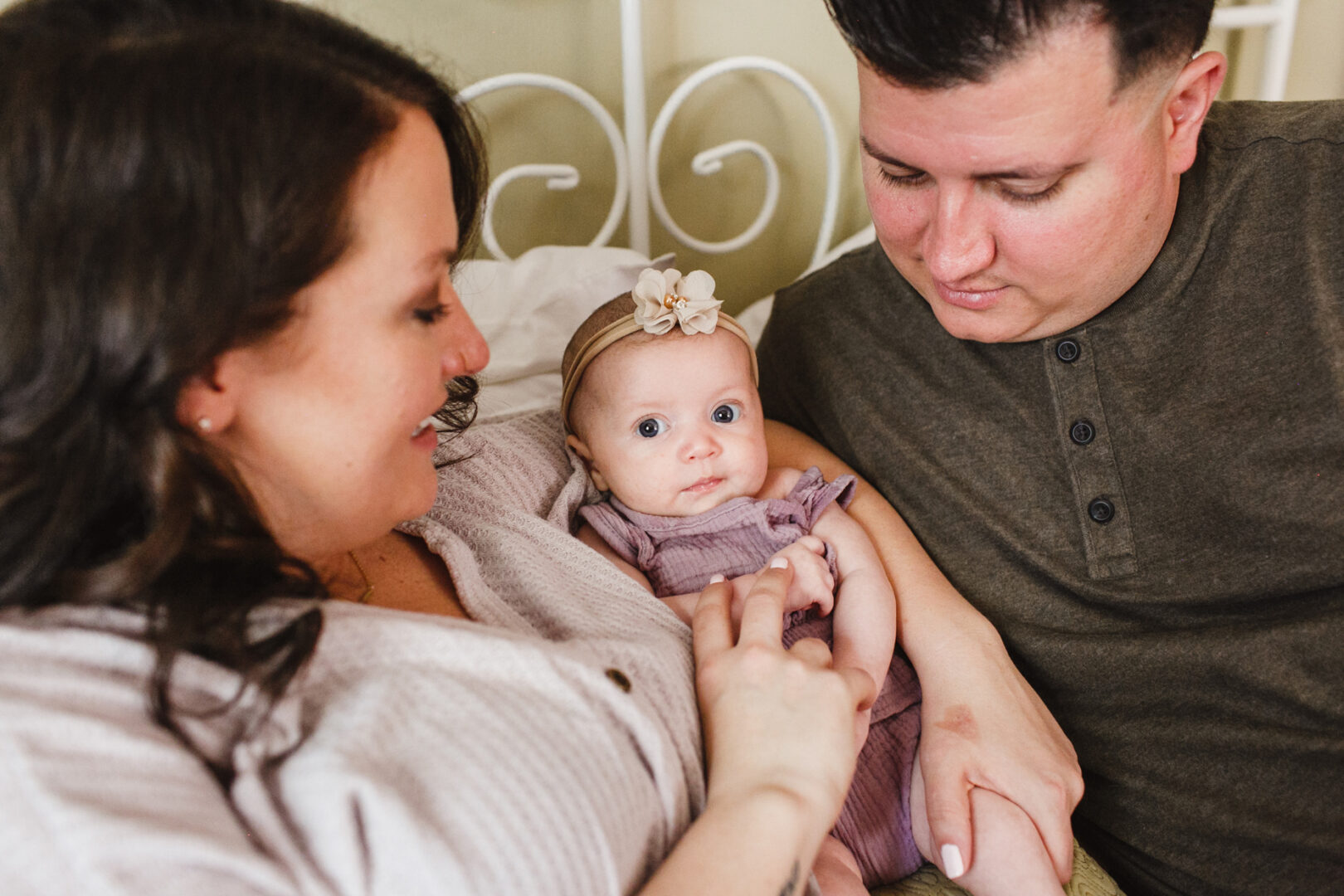 A baby wearing a floral headband is held by two adults, sitting closely on a bed as they admire the newborn. This tender moment captures the essence of newborn photos in Philadelphia.