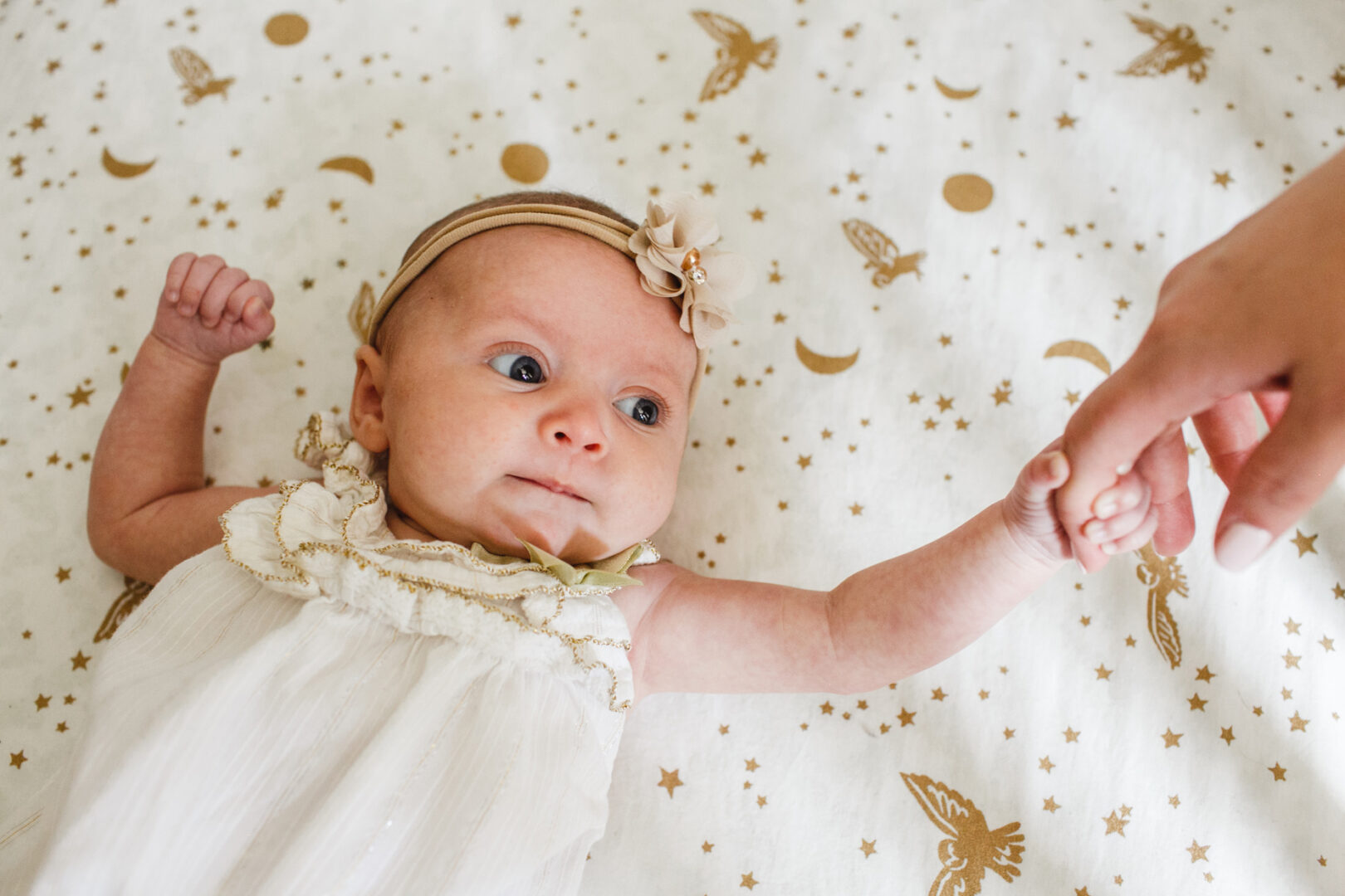 A newborn in a white dress and headband lies on a star-patterned blanket, gently clutching an adult's finger. Perfect for capturing those precious moments, this scene is an ideal addition to any collection of newborn photos in Philadelphia.