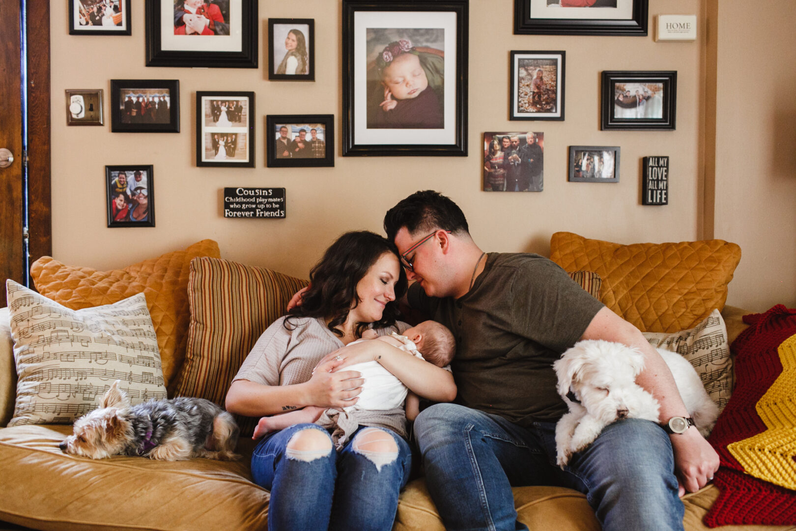 A couple sits on a sofa with their baby, surrounded by beautifully framed newborn photos of Philadelphia adorning the wall. Two small dogs snuggle close by, completing the charming family scene.