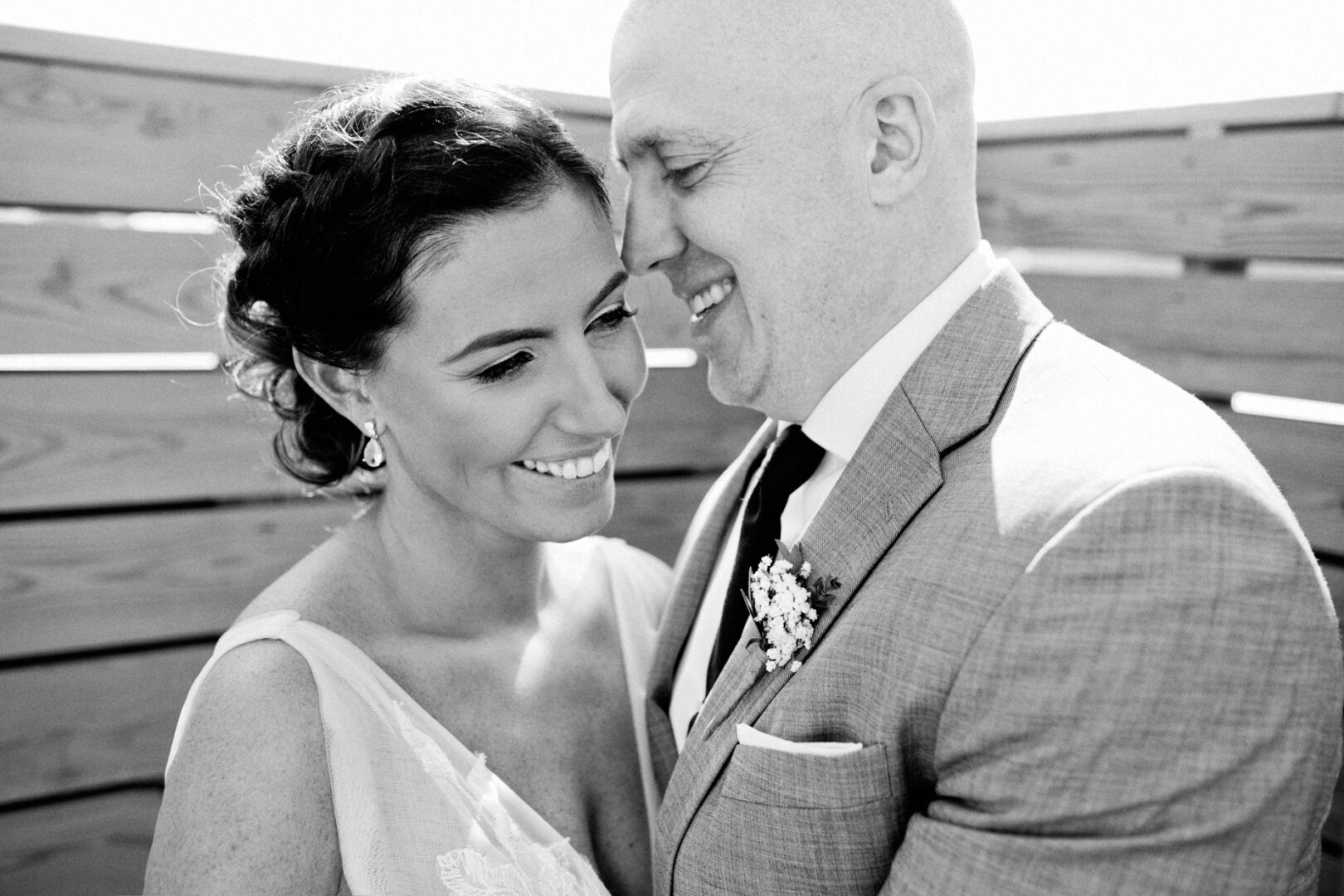 A couple in formal attire happily embracing, standing in front of a wooden fence near Seaport Pier, with the man wearing a light-colored suit and boutonniere. Black and white photo.