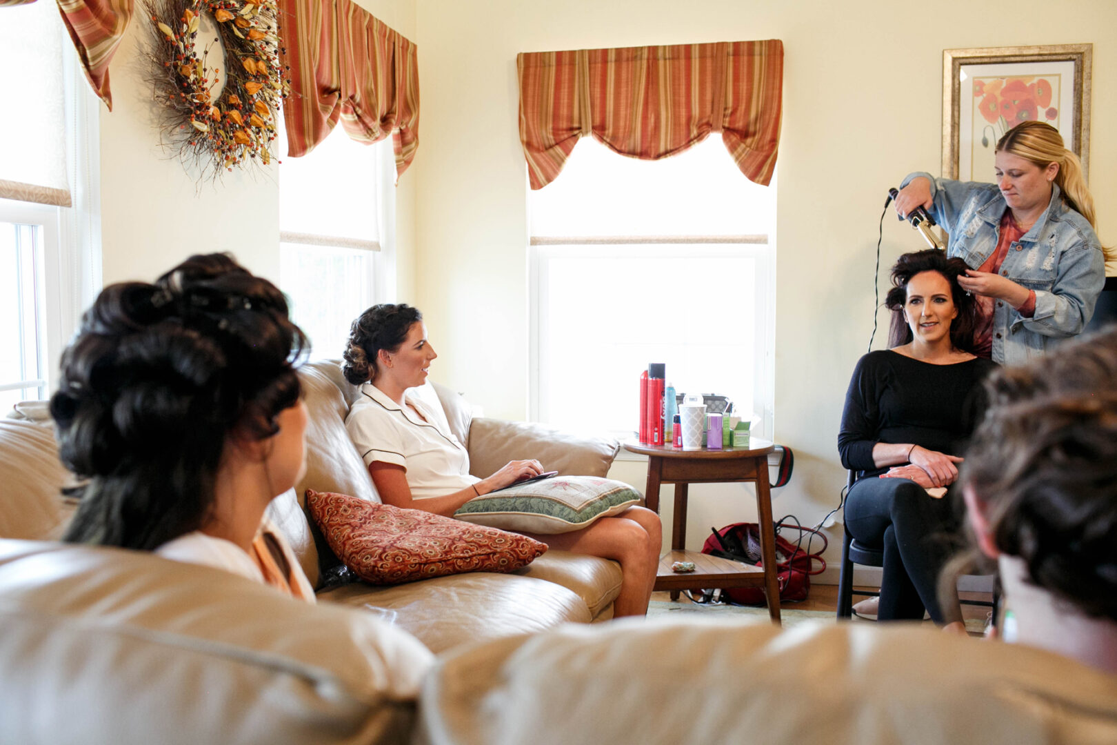 Two women sit on a couch in a cozy living room, while another styles a third woman's hair. The space, reminiscent of a Seaport Pier ambience, features a wreath, striped curtains, and a painting on the wall.