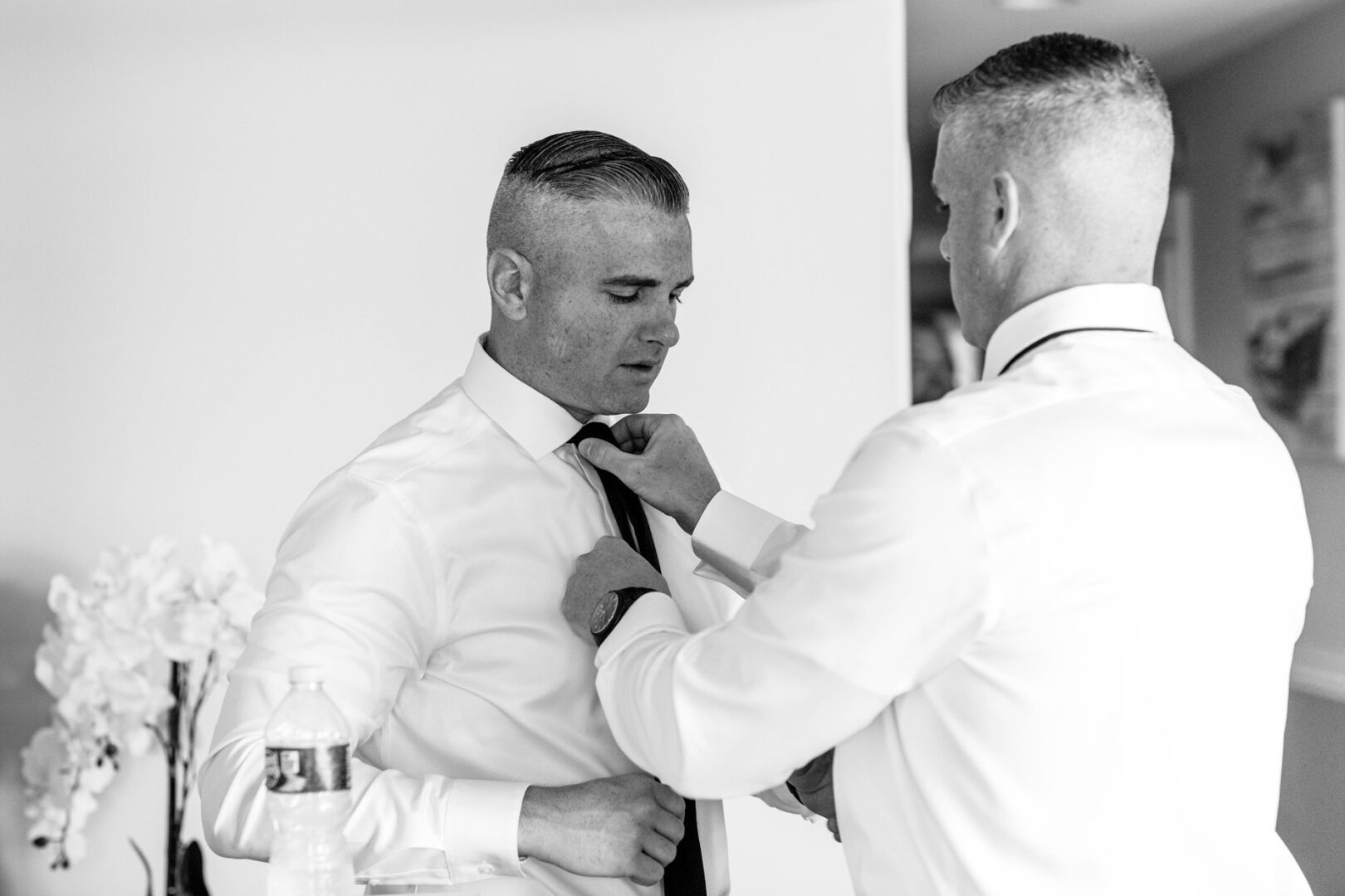 In a black and white image, two men in white shirts stand in a room reminiscent of a cozy spot near Seaport Pier. One adjusts the other's tie while a flower and a water bottle sit elegantly on the table.