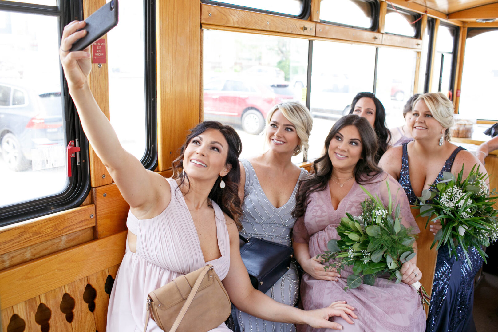 Five women in dresses take a selfie together on a wooden trolley near Seaport Pier, some holding green bouquets.