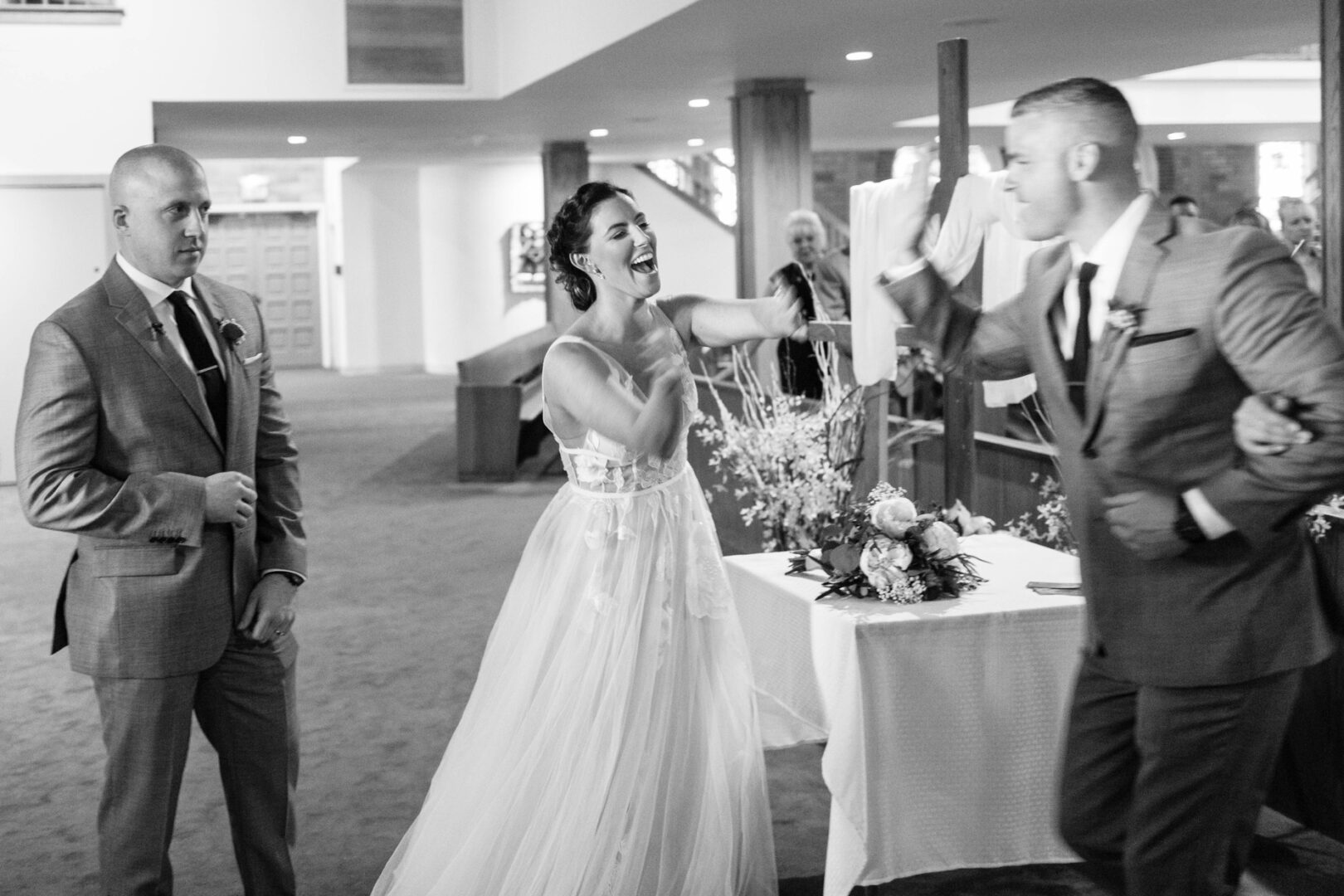 At Seaport Pier, the bride playfully interacts with the groom at a wedding surrounded by guests. Two men in suits stand nearby, adding to the lively scene, while a beautifully decorated table with flowers completes the elegant indoor setting.