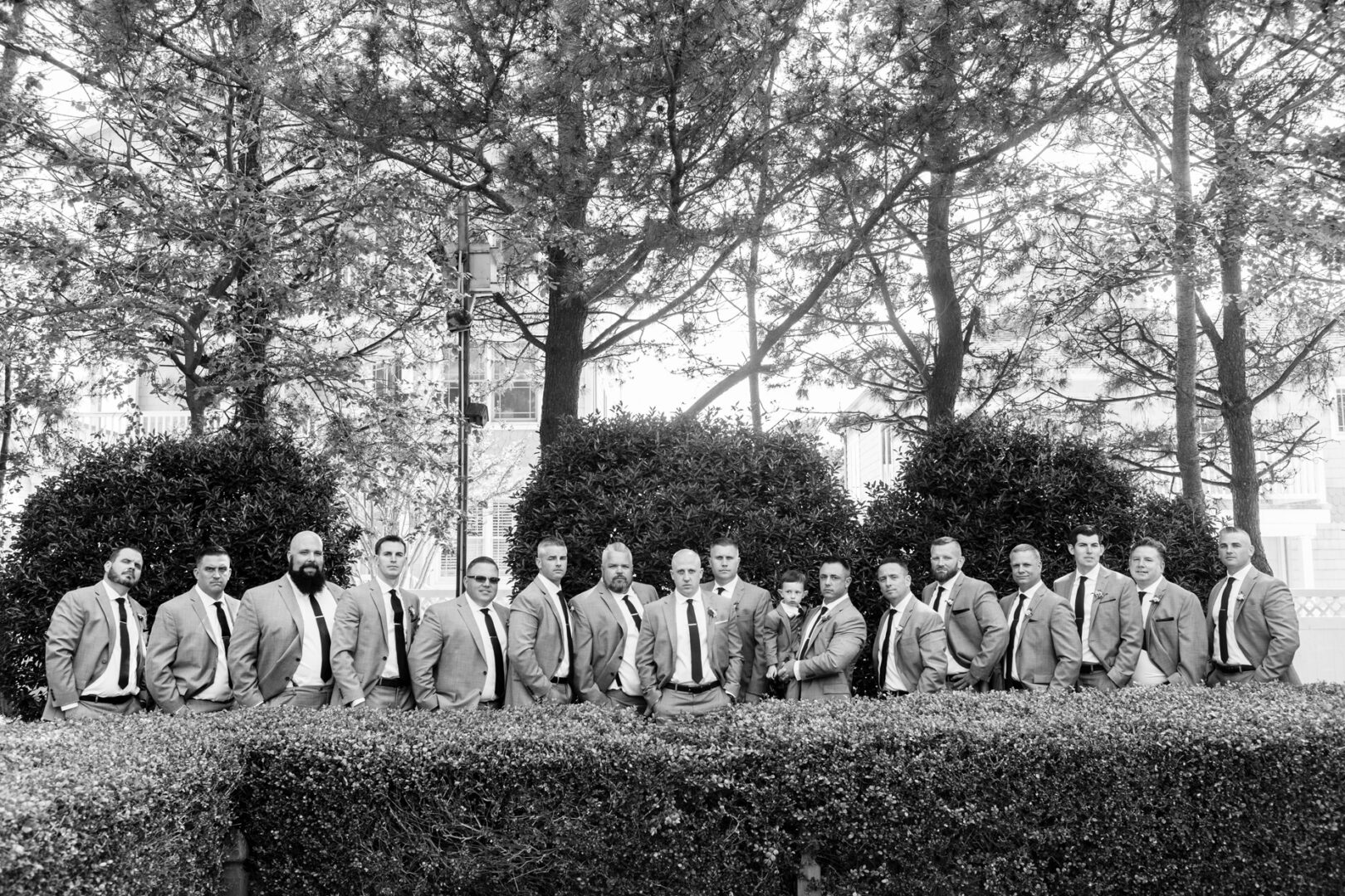 A group of men in suits and ties stand in a row behind a hedge, with trees reminiscent of those you might find at Seaport Pier. The image is elegantly captured in black and white.