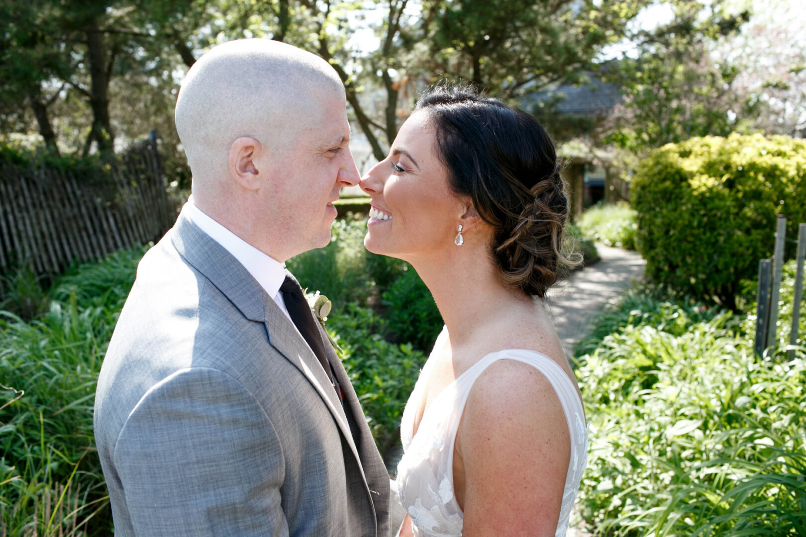 A bride and groom face each other outdoors, smiling amidst the lush greenery and trees. The groom wears a gray suit, and the bride is stunning in her white dress. Just beyond them, the serene backdrop of Seaport Pier adds a touch of coastal charm to their special moment.