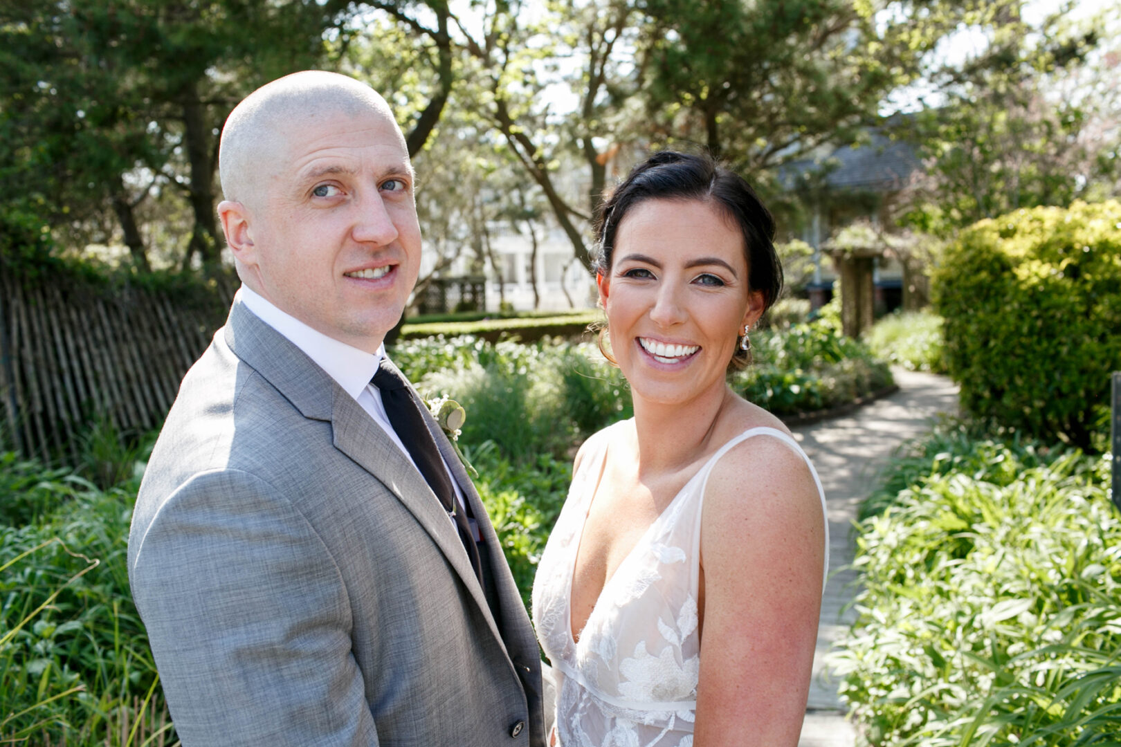 A smiling couple, one in a suit and the other in a lace dress, poses together on a garden pathway with trees and shrubs reminiscent of the serene views at Seaport Pier.
