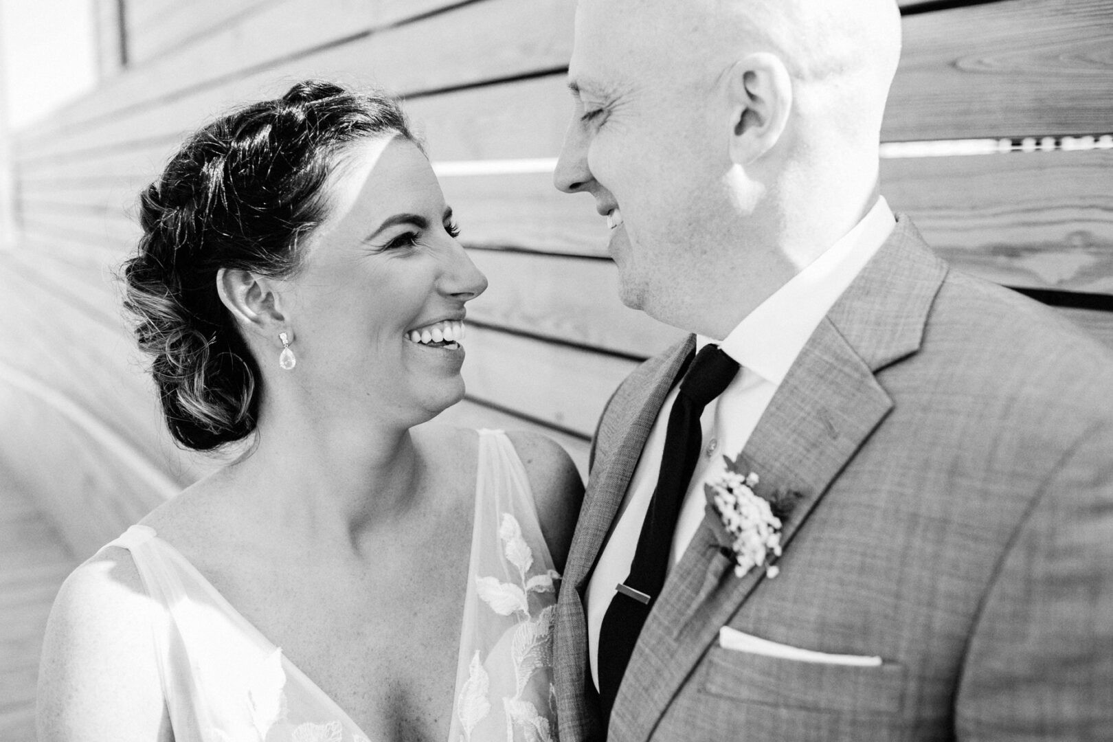 A smiling couple, dressed formally, stand closely together in front of a wooden wall at Seaport Pier. The woman wears earrings, and the man has a boutonniere on his suit. Black and white image.