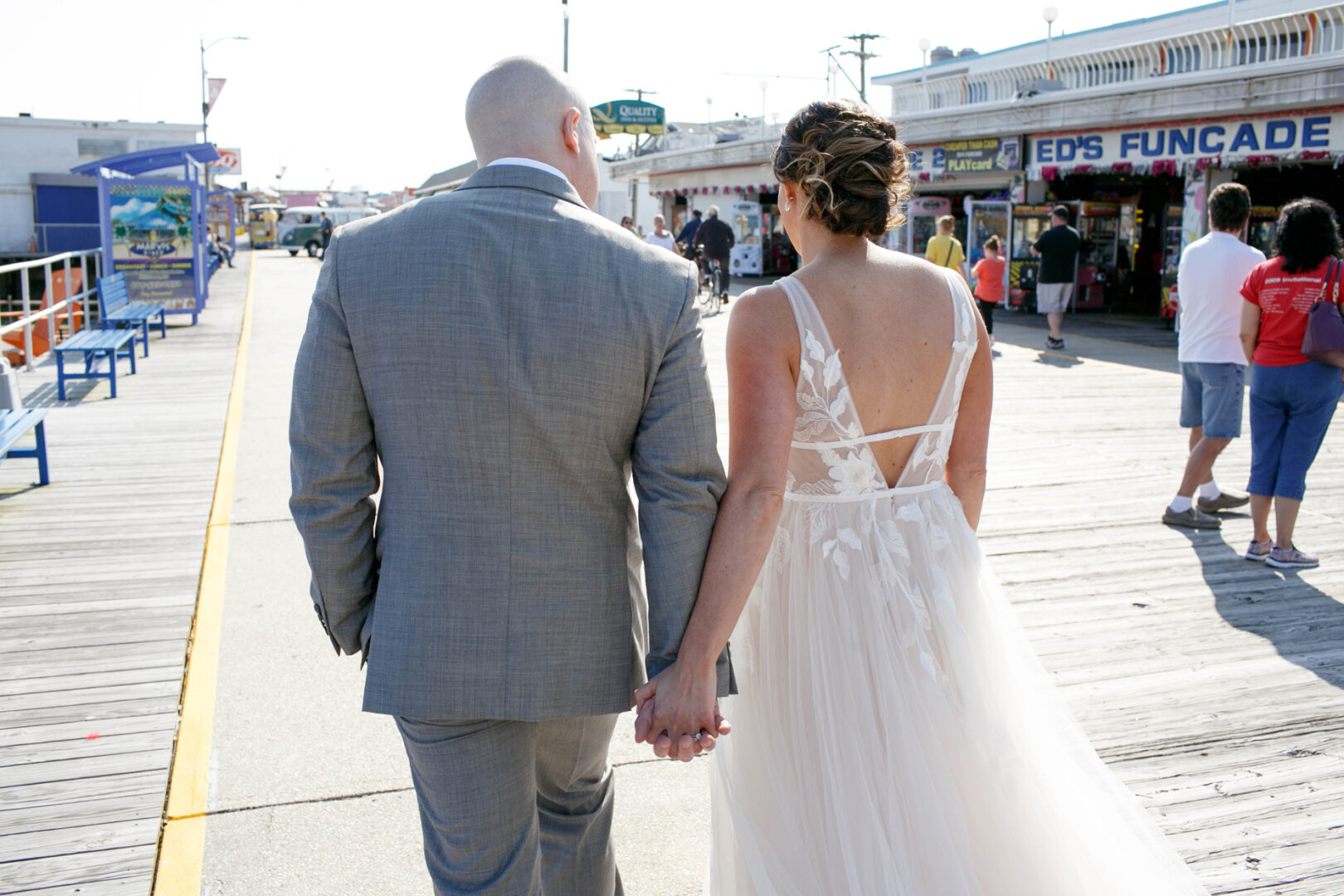 A couple in formal attire strolls hand-in-hand along the Seaport Pier boardwalk, with shops and people bustling in the background.