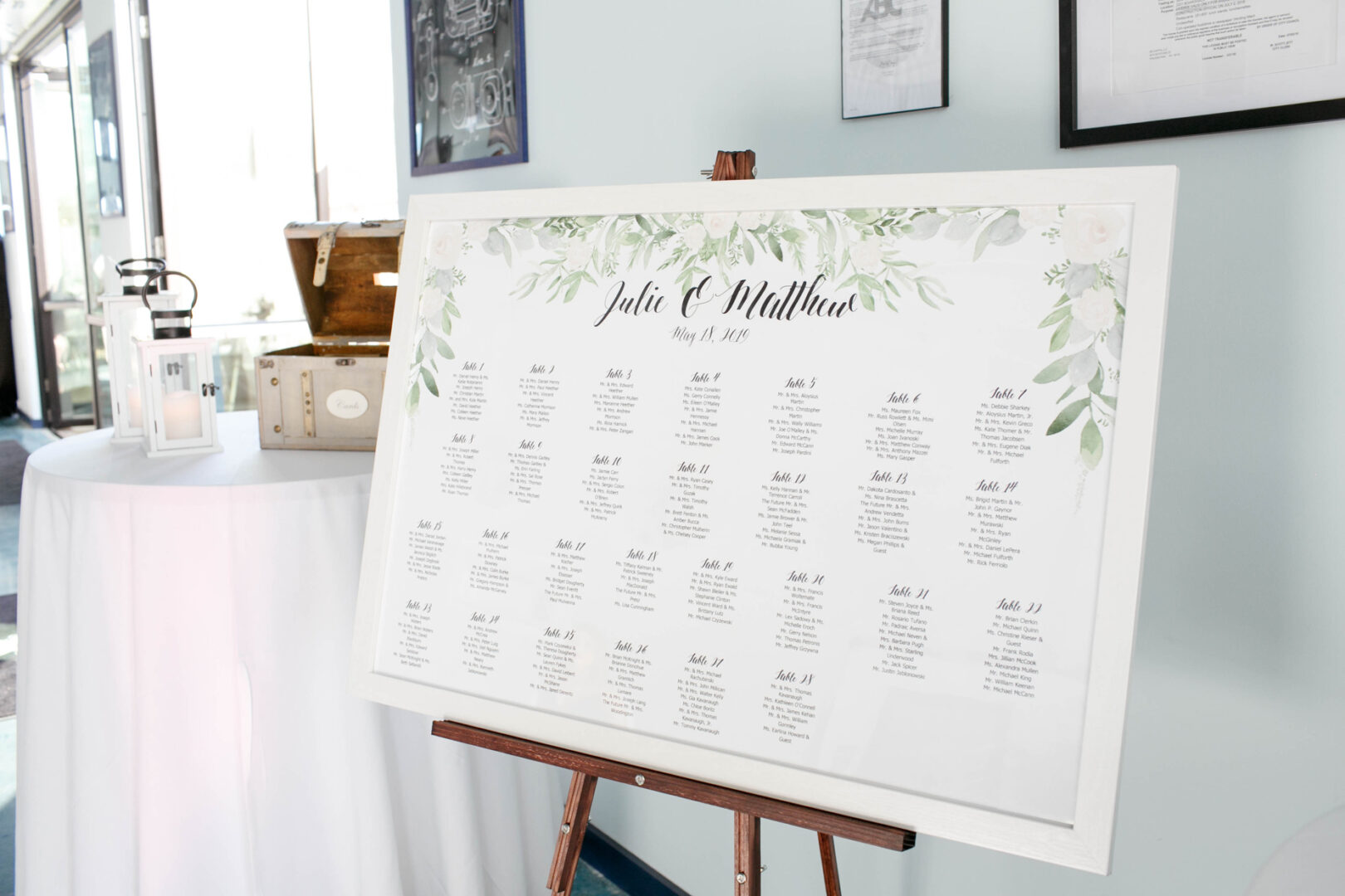 A wedding seating chart on an easel, adorned with greenery and floral designs, lists tables and guest names. Nearby, a wooden box and a lantern evoke the charm of Seaport Pier, setting a picturesque scene for guests to admire.