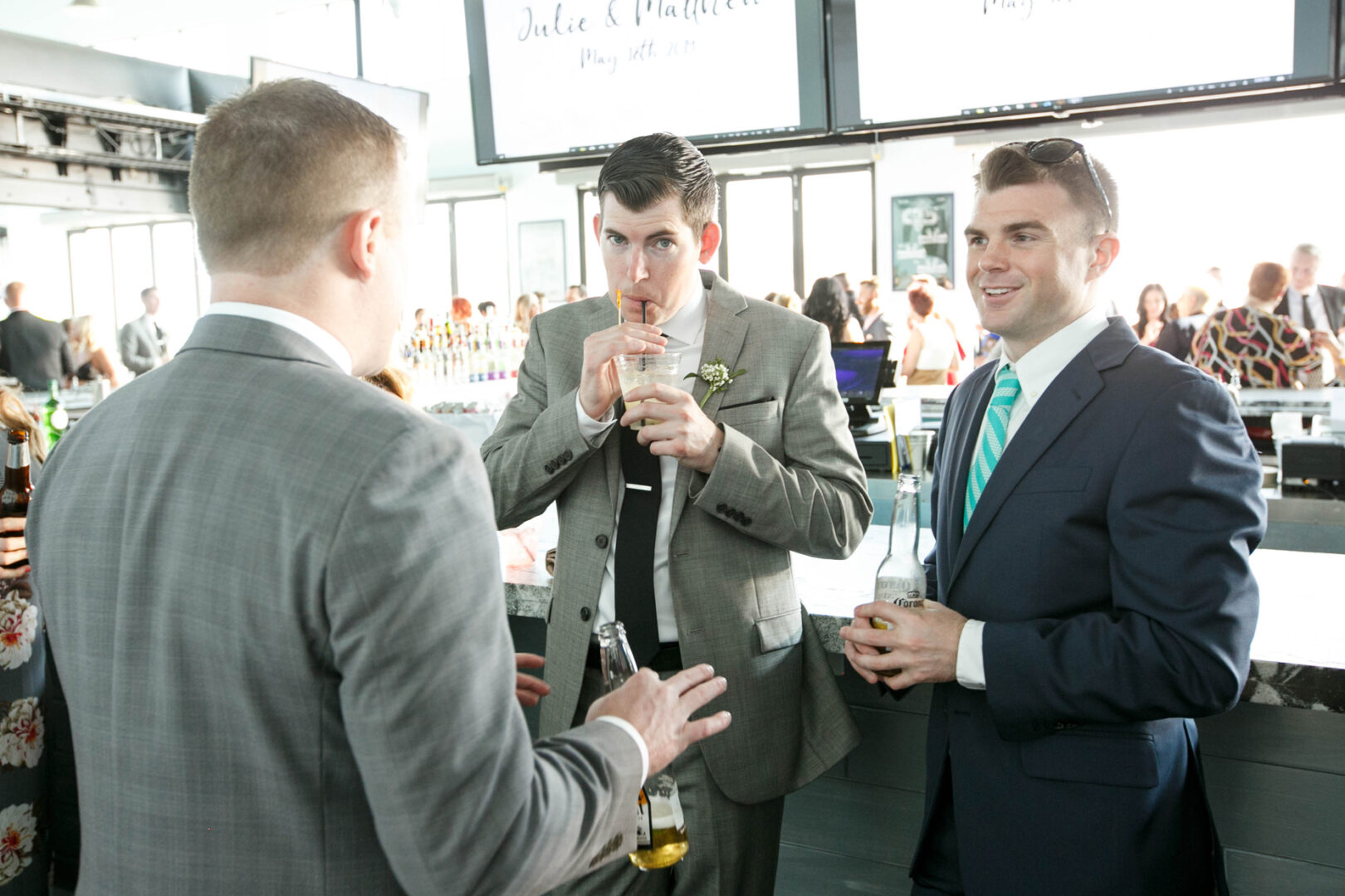 At Seaport Pier, three men in suits convene at a bar during a lively social event. Two hold drinks, one casually sipping from a straw, as the vibrant crowd buzzes in the background.