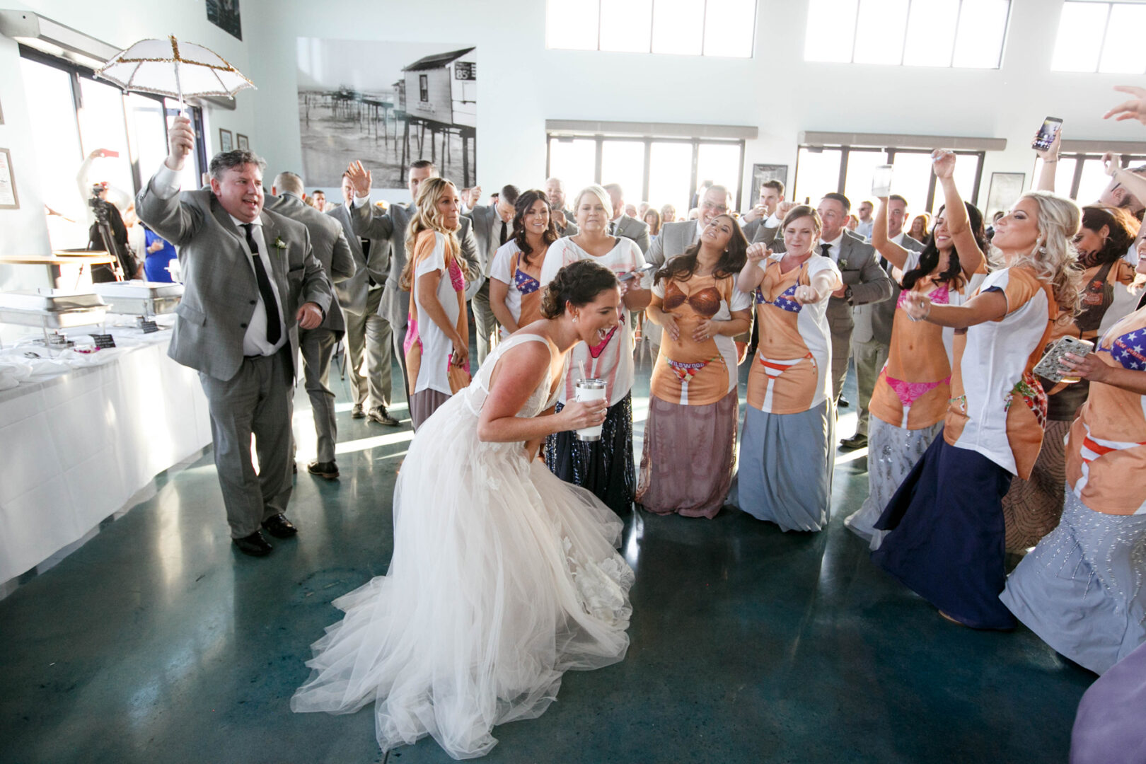 The bride danced joyfully at the lively wedding reception, set against the backdrop of Seaport Pier. Cheering adults, dressed in formal wear, casual outfits, and even some in swimwear, surrounded her amidst the bright windows and white walls.
