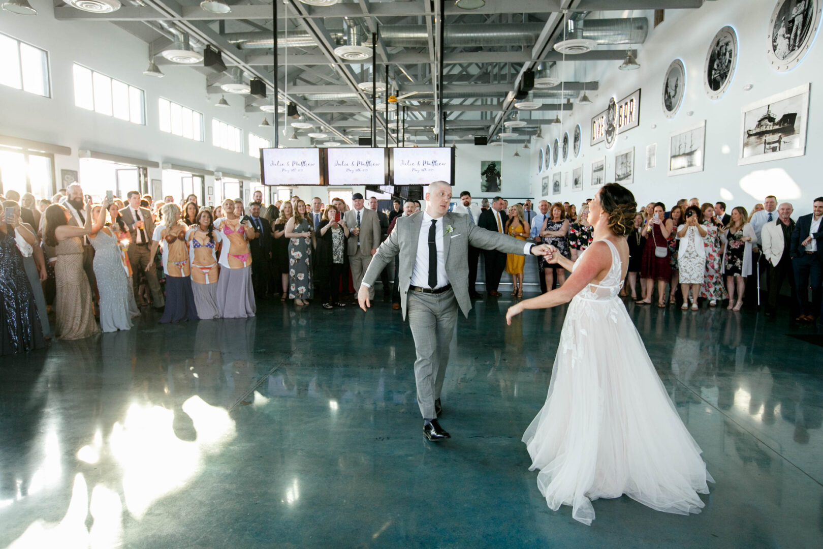 A couple dances gracefully at Seaport Pier, the venue's shiny floor reflecting their every step. The groom, in a distinguished gray suit, leads the bride, whose flowing white dress catches the light streaming through expansive windows as the audience looks on in admiration.