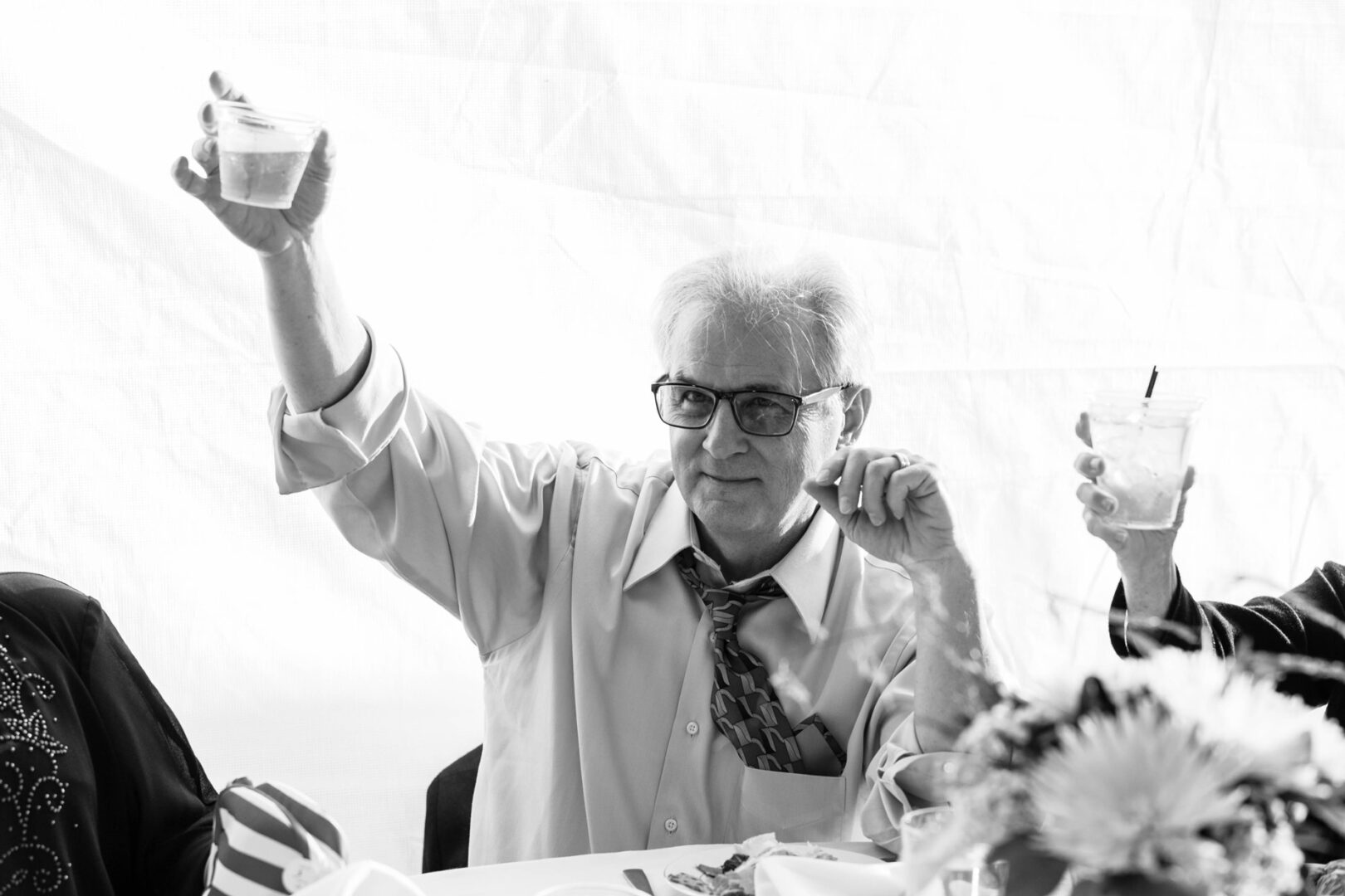 An older man in glasses and a loosened tie raises a drink at a gathering near Seaport Pier, with a festive table setting in front of him.