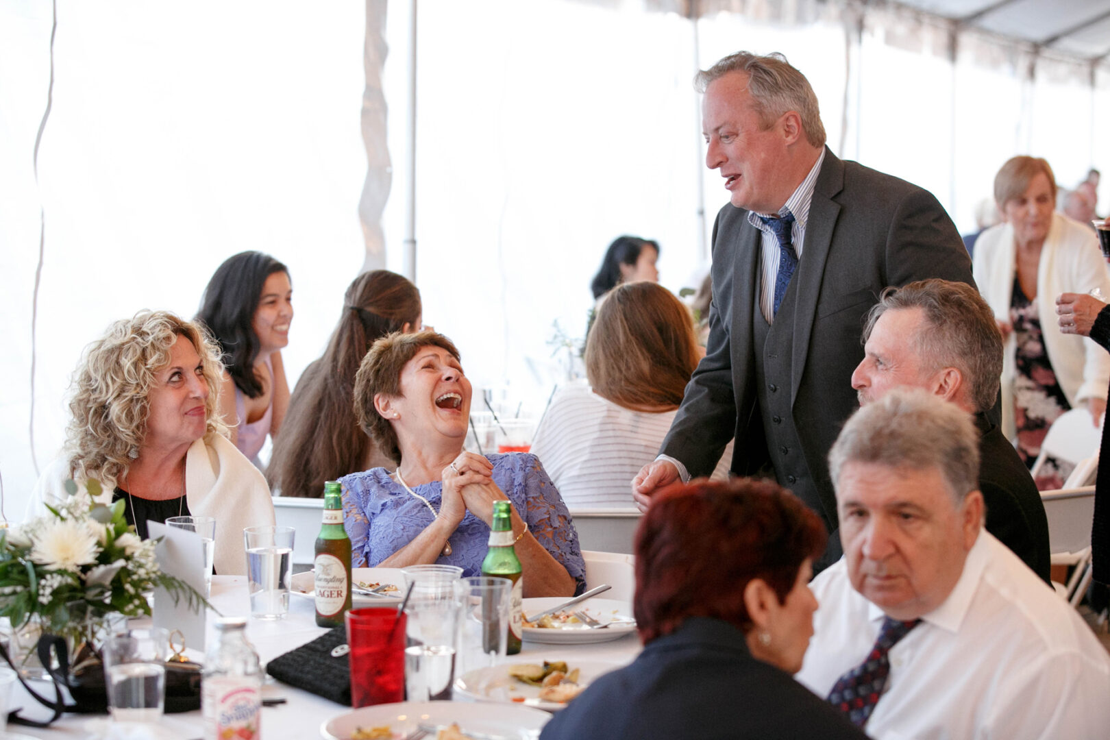 People seated around a table at Seaport Pier, enjoying plates of food and drinks. A man in a suit stands, engaging with others, while a woman bursts into laughter.