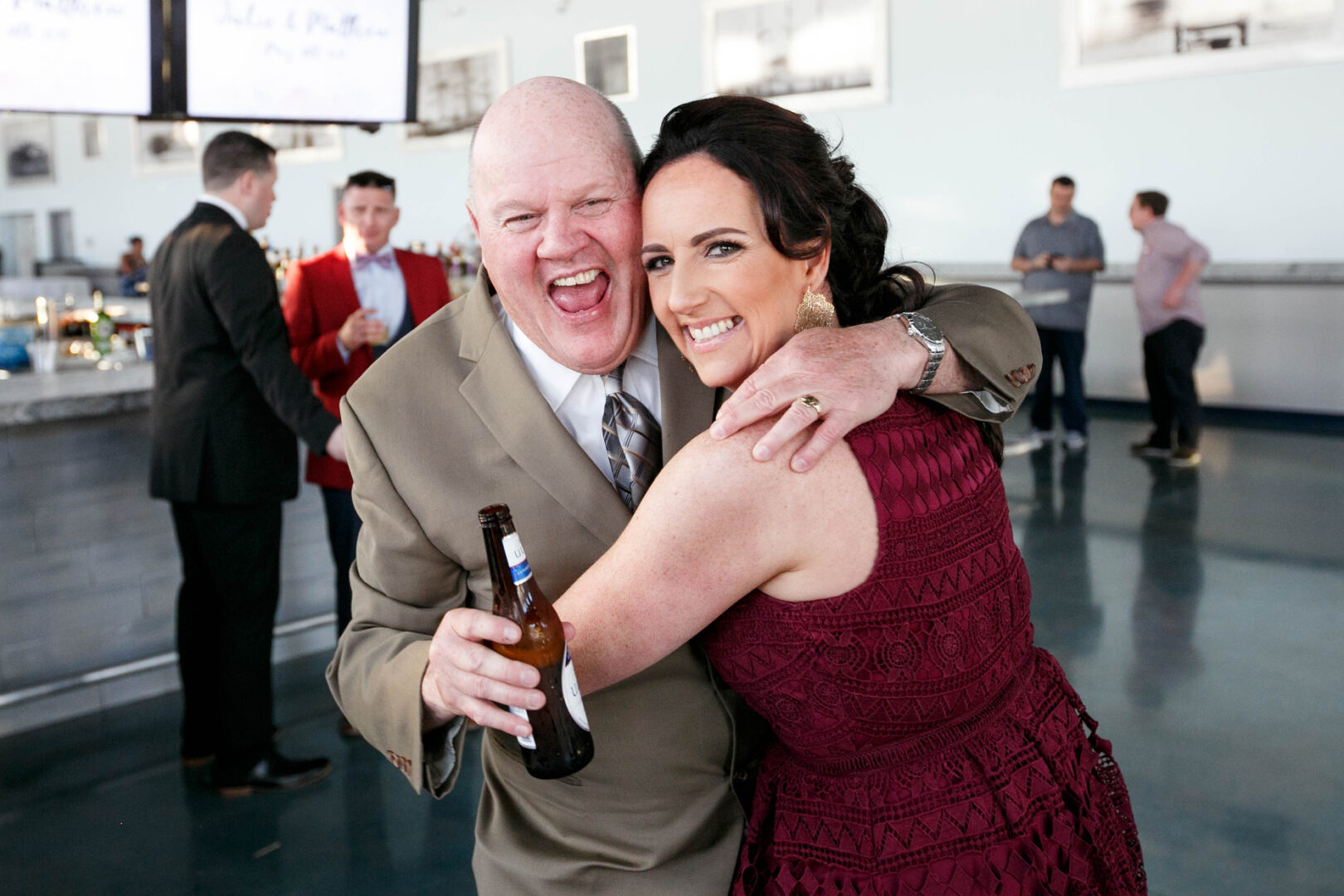 Man in a beige suit smiling, holding a beer, with a woman in a burgundy dress hugging him at Seaport Pier. Other people and a bustling bar are in the background.