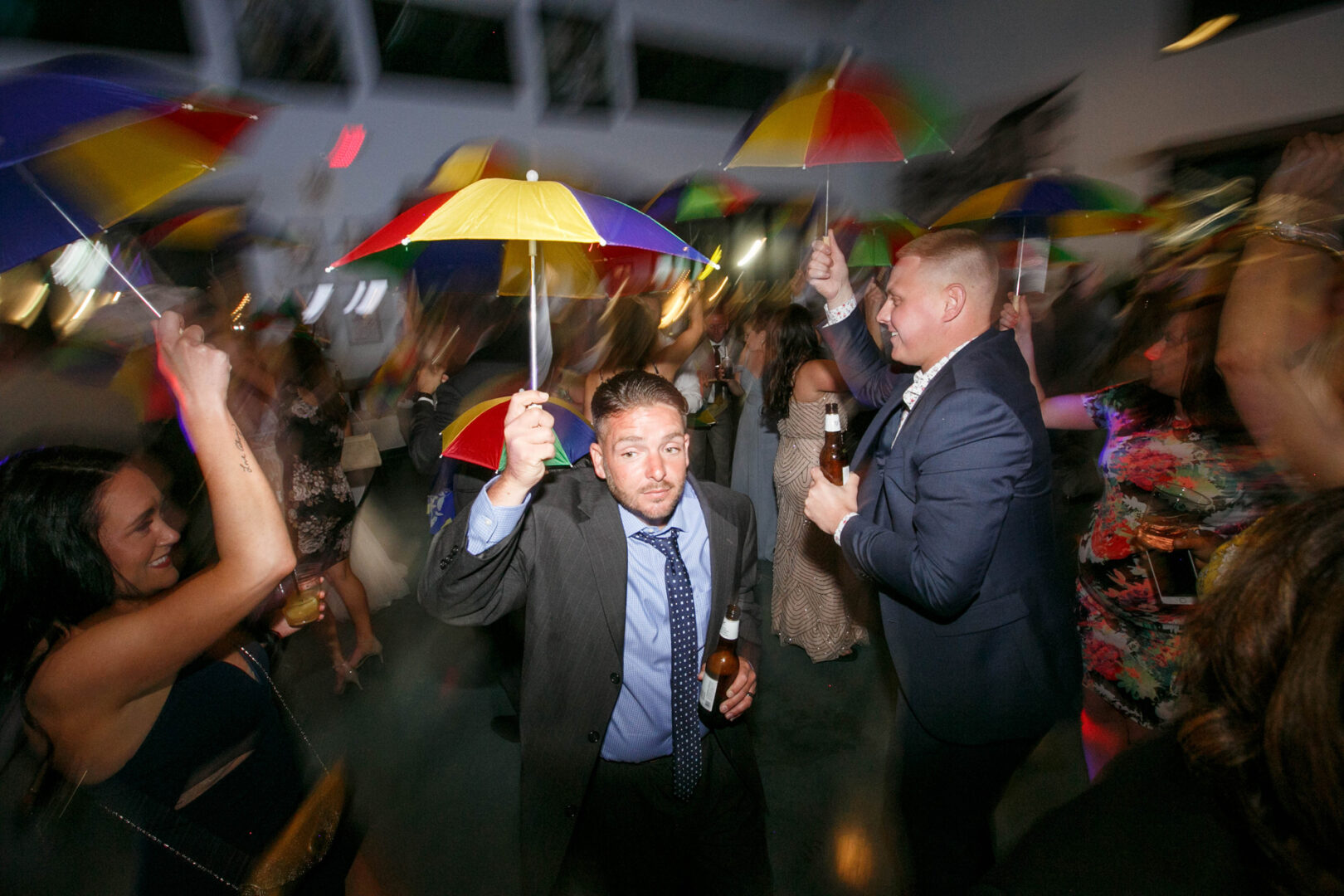 Under the vibrant lights of Seaport Pier, people with colorful umbrellas fill the dance floor, some clutching drinks in hand. The motion blur captures their energetic movements, creating a lively and unforgettable atmosphere by the water's edge.