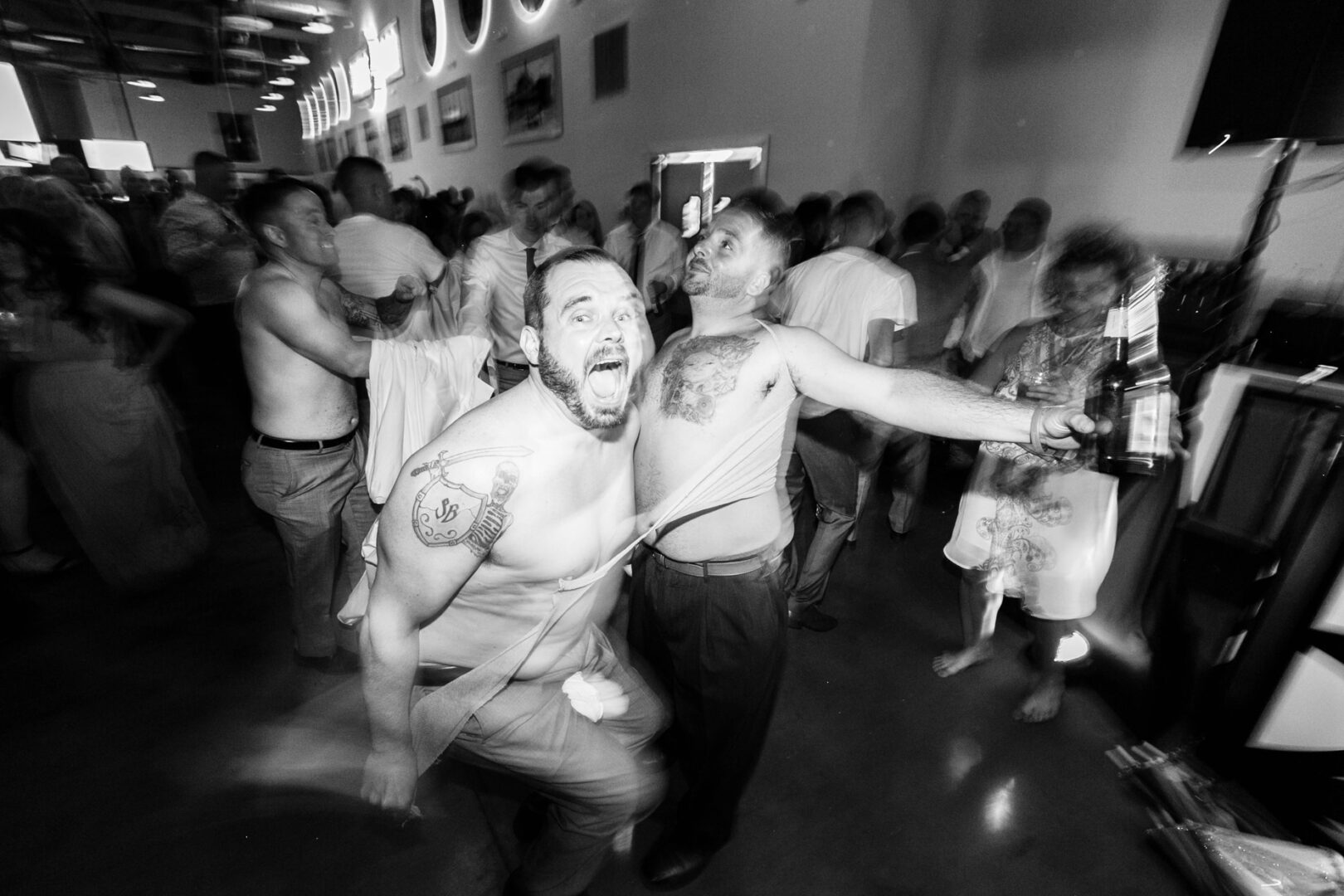 Two shirtless men energetically dance amidst a lively crowd at Seaport Pier, with one holding a beer bottle, capturing the vibrant atmosphere of the indoor event.