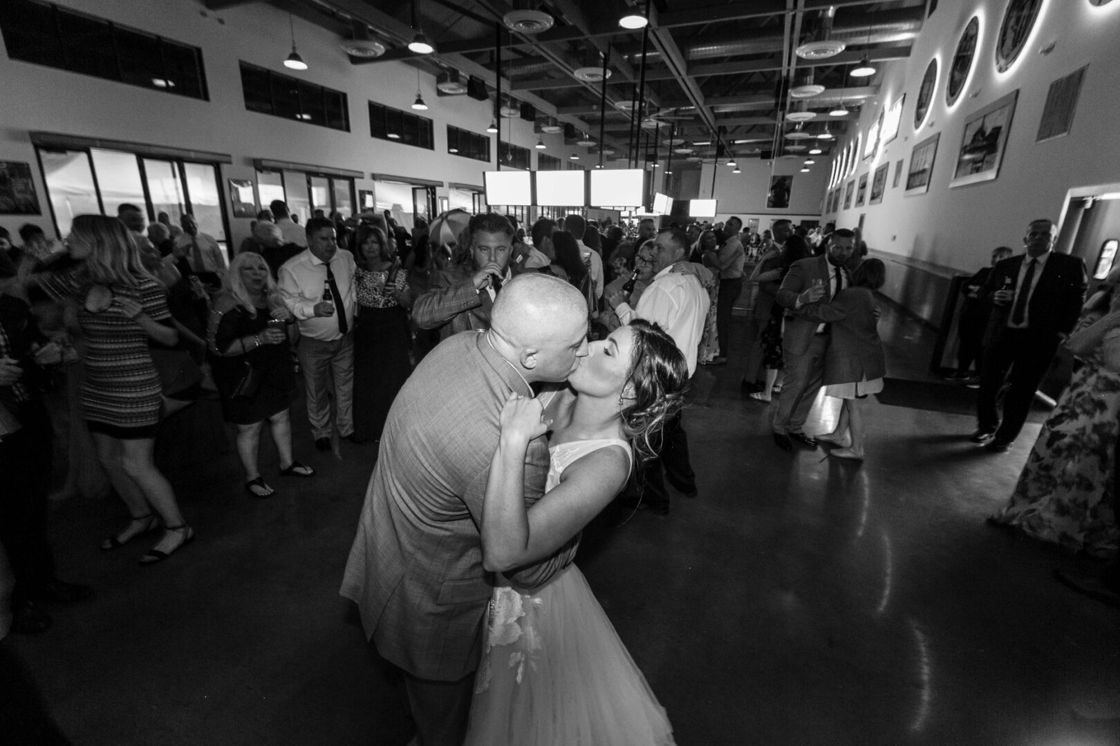 Amidst the lively ambiance of Seaport Pier, a bride and groom share a tender kiss on the dance floor, surrounded by delighted guests in the expansive hall.
