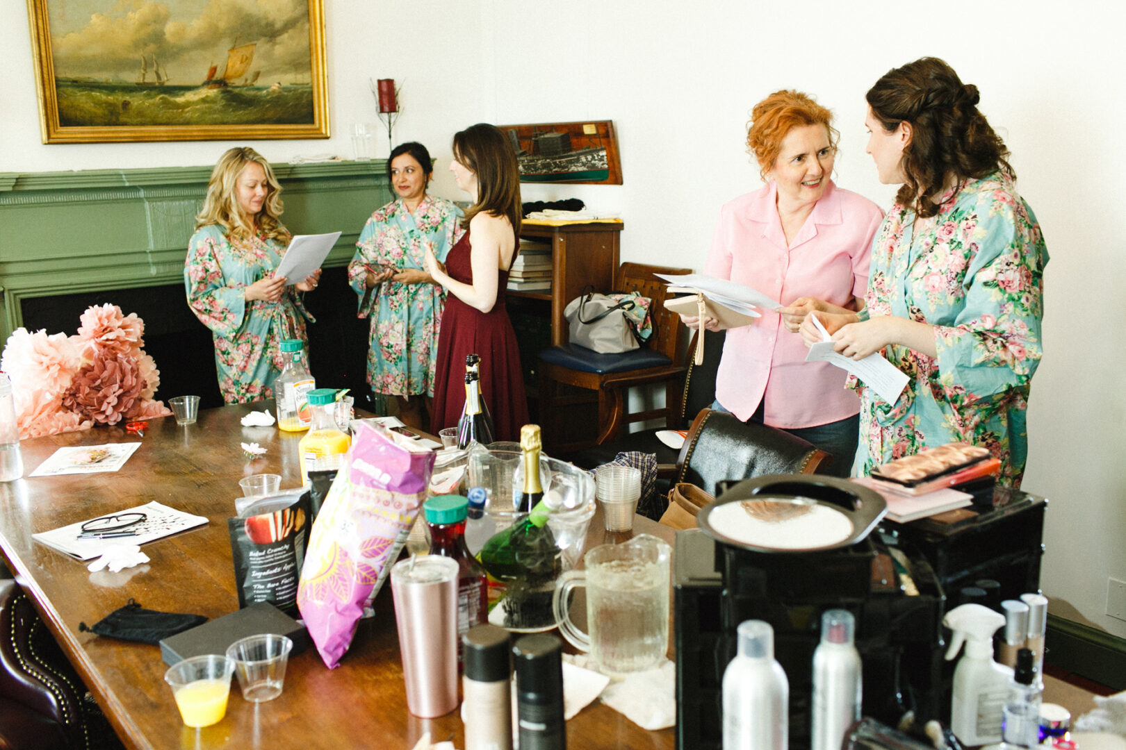 Five women gather in a room at the Corinthian Yacht Club of Philadelphia, where a table brims with drinks and snacks. Four don floral robes, while one stands out in a red dress. They examine papers closely, preparing for the day's elegant wedding event.