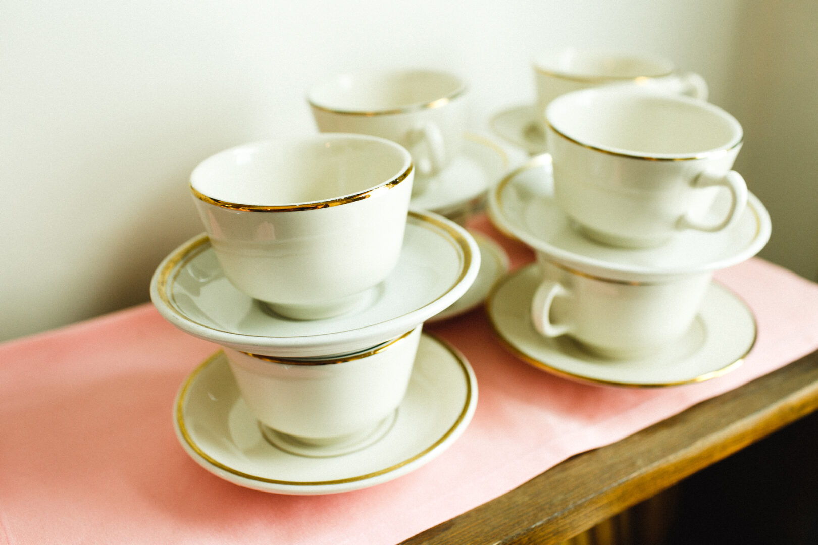 Five white teacups with saucers, each edged with a gold rim, are elegantly arranged on a wooden surface adorned with a pink cloth—perfect for adding a touch of grace to your Corinthian Yacht Club of Philadelphia wedding setting.