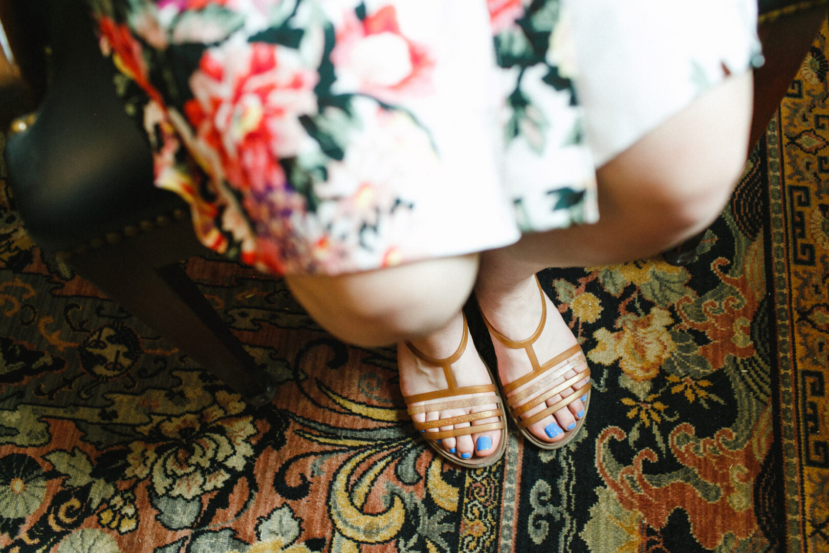 A person, dressed in a floral dress and tan sandals with blue nail polish, sits gracefully on a patterned rug, evoking the elegance of a Corinthian Yacht Club of Philadelphia wedding.
