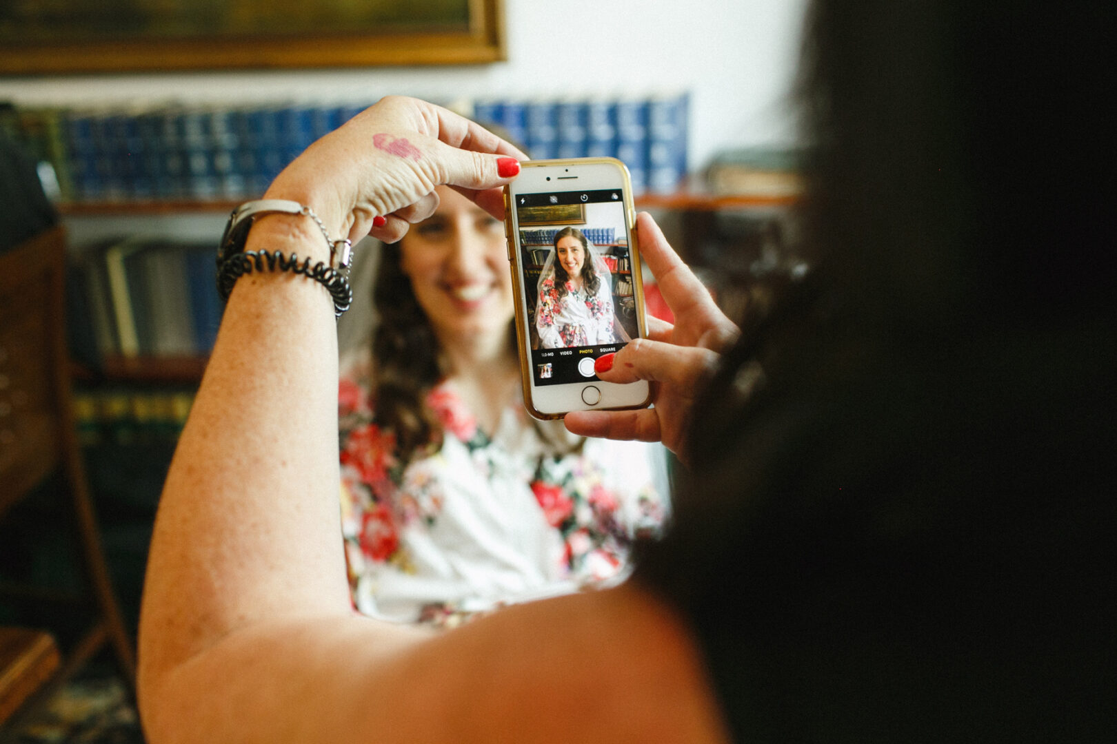 A person captures a photo of a woman, whose smile shines as brightly as a Corinthian Yacht Club of Philadelphia wedding. She's wearing a floral top, with shelves brimming with books subtly visible in the background.