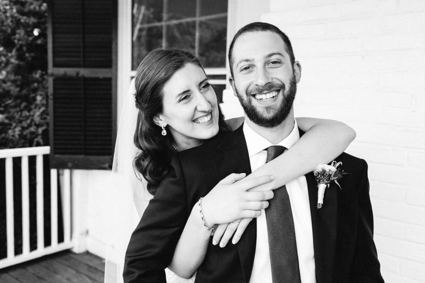 A smiling couple poses together on a porch at their Corinthian Yacht Club of Philadelphia wedding; the woman hugs the man from behind. Both are dressed in formal attire in this enchanting black and white image.